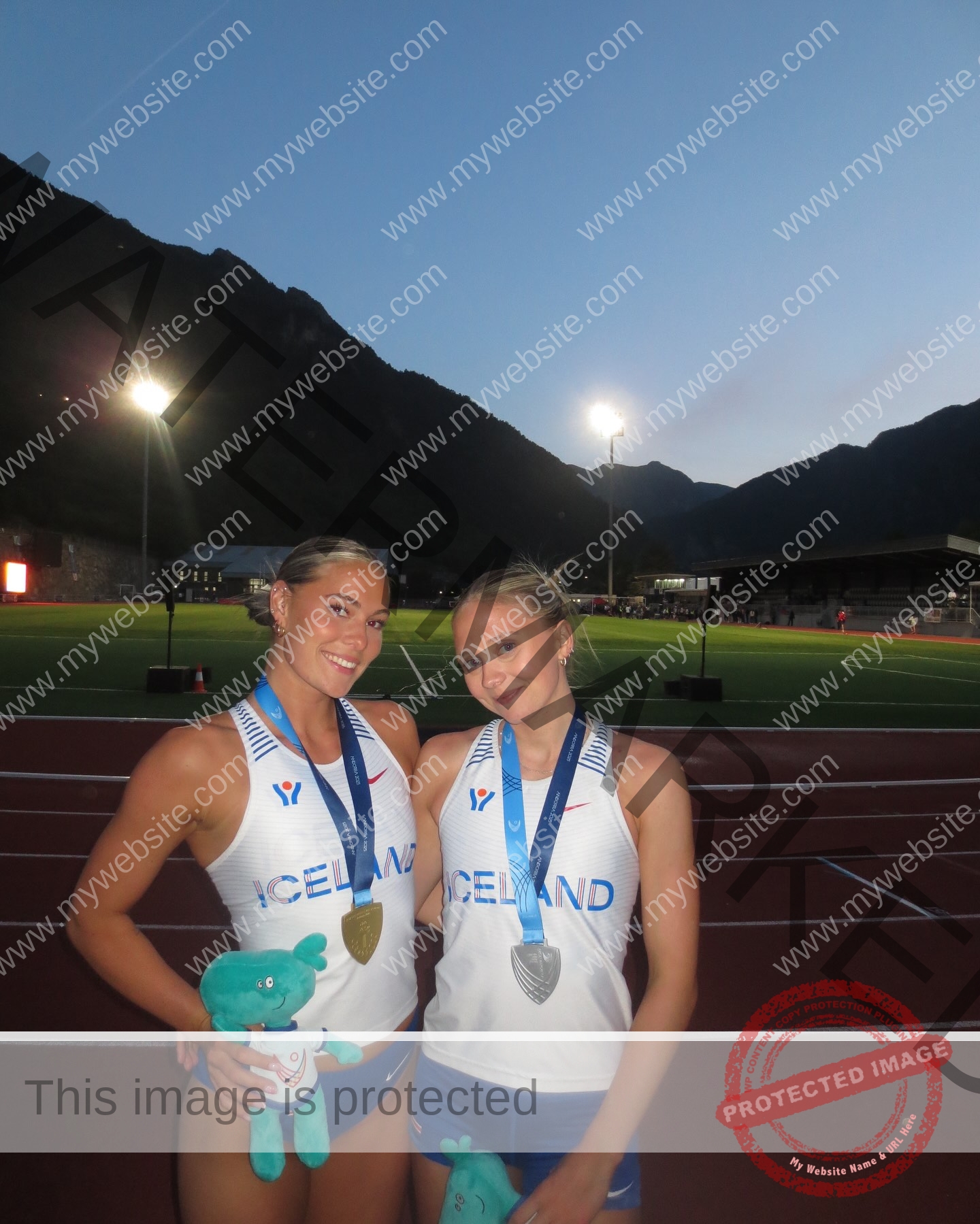birna-kristjansdottir-iceland-birnakristink-11052 Birna-Kristjansdottir, track athlete from Iceland, stands at dusk on a track with another athlete, both holding medals and mascots.