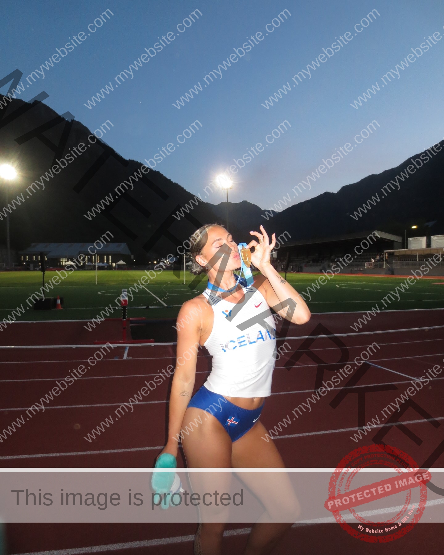 birna-kristjansdottir-iceland-birnakristink-11054 Birna-Kristjansdottir, track athlete from Iceland, stands on a track at dusk with a medal in her mouth and shoes in hand.