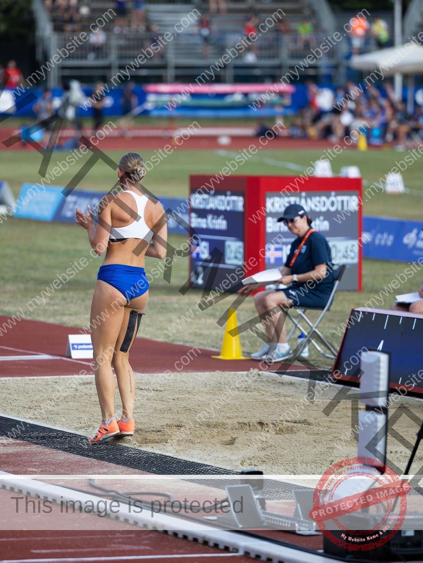 birna-kristjansdottir-iceland-birnakristink-11066 Birna-Kristjansdottir, track athlete from Iceland, in a white top and blue shorts prepares to long jump; judge and crowd nearby.