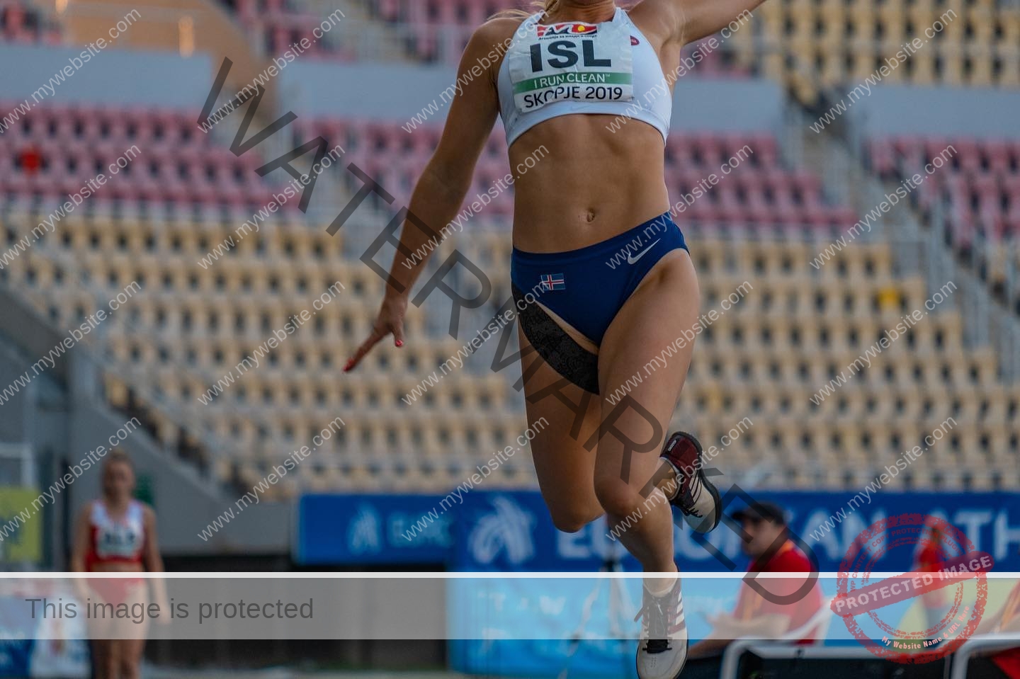 birna-kristjansdottir-iceland-birnakristink-11095 Birna-Kristjansdottir, track athlete from Iceland, leaps mid-air in long jump at a stadium with ISL bib; seats and staff behind.