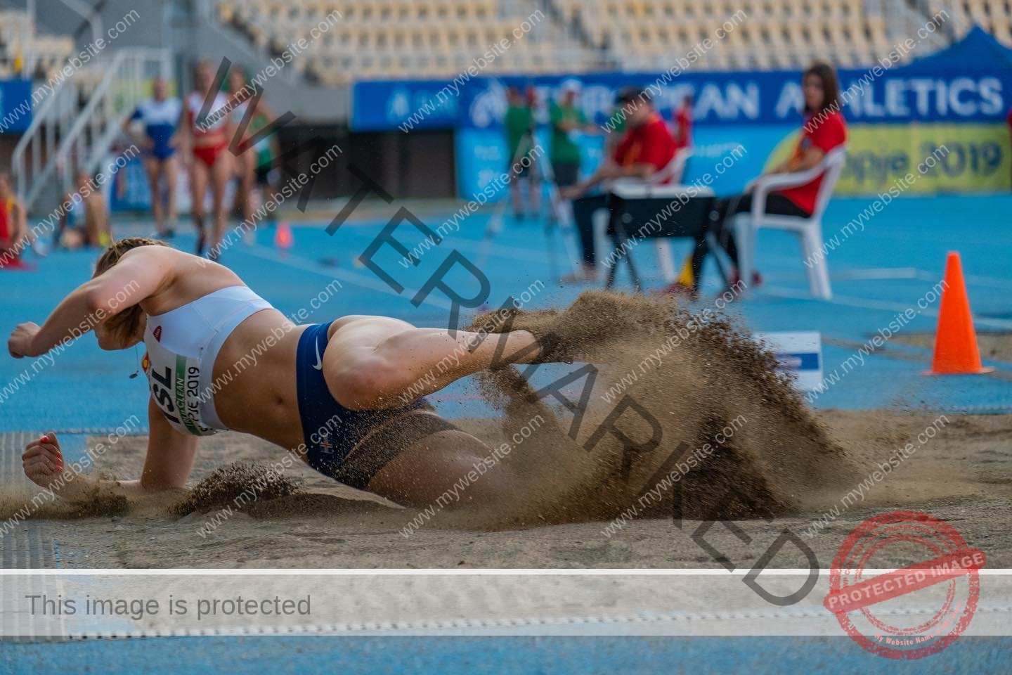 birna-kristjansdottir-iceland-birnakristink-11097 Birna-Kristjansdottir, track athlete from Iceland lands in a sandpit during long jump, sand flying as others watch on the blue track.