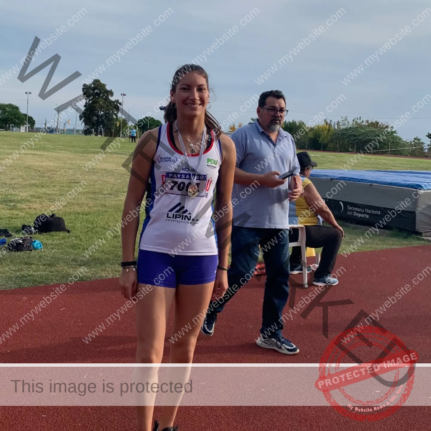 brandy-romero-uruguay-brandy_rom08-0004 Brandy Romero Brandy Romero, a female track and field athlete from Uruguay with a medal and running bib, smiles on the track. A man with a phone stands behind her amidst sports gear and grassy fields.