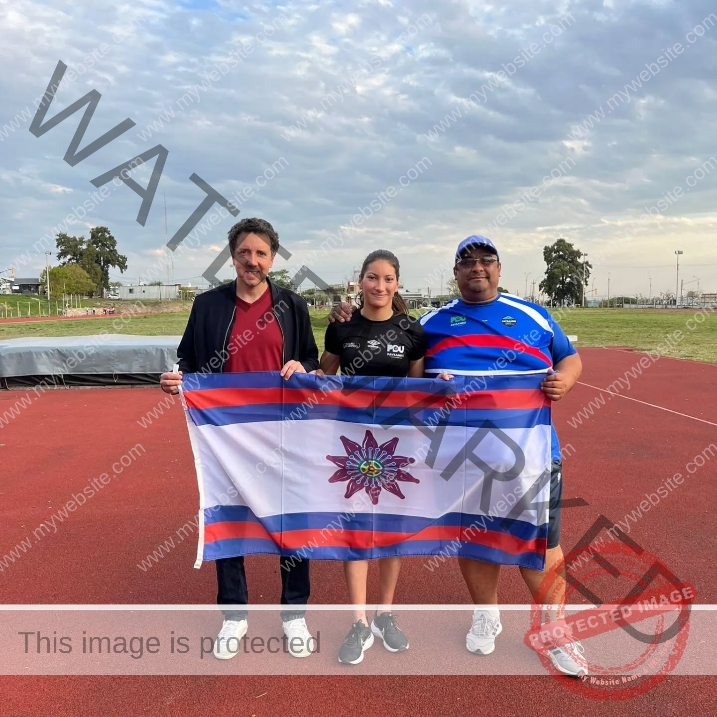 brandy-romero-uruguay-brandy_rom08-0011 Brandy Romero Three people stand on a red athletics track holding a white flag with blue and red stripes and a floral emblem; cloudy sky.