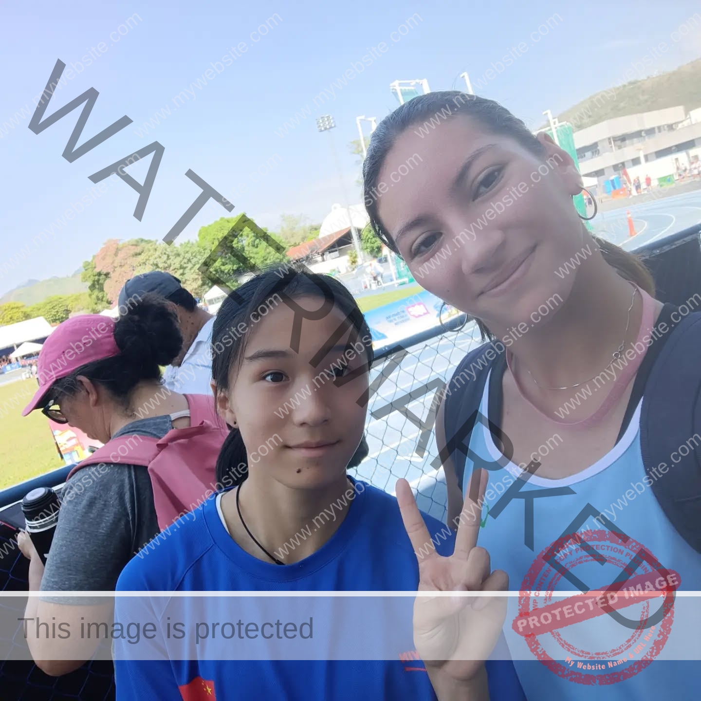 brandy-romero-uruguay-brandy_rom08-0018 Brandy Romero Two young women stand together near a track in Uruguay, smiling; one does a peace sign. Sunny sports area in the background.