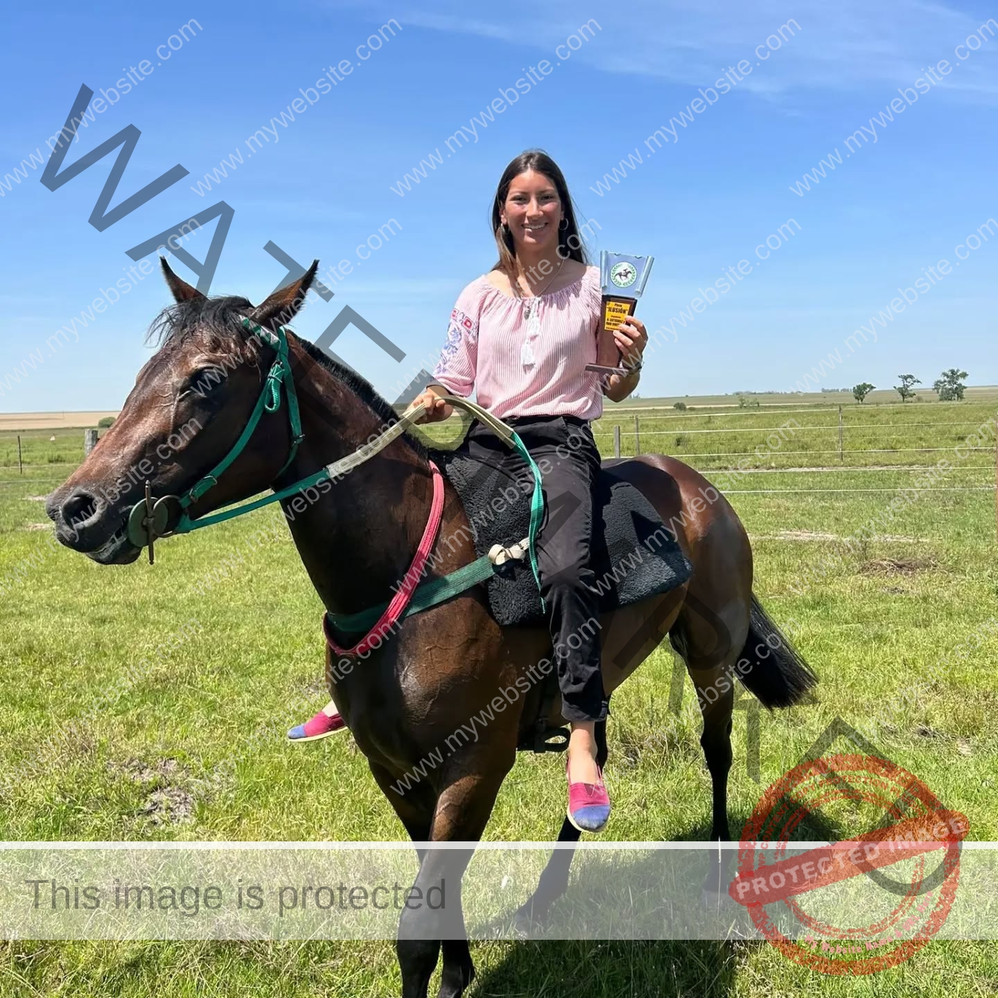 brandy-romero-uruguay-brandy_rom08-0031 Brandy Romero Brandy Romero, a smiling woman from Uruguay, sits on a brown horse in a grassy field, holding a trophy. She wears pink.
