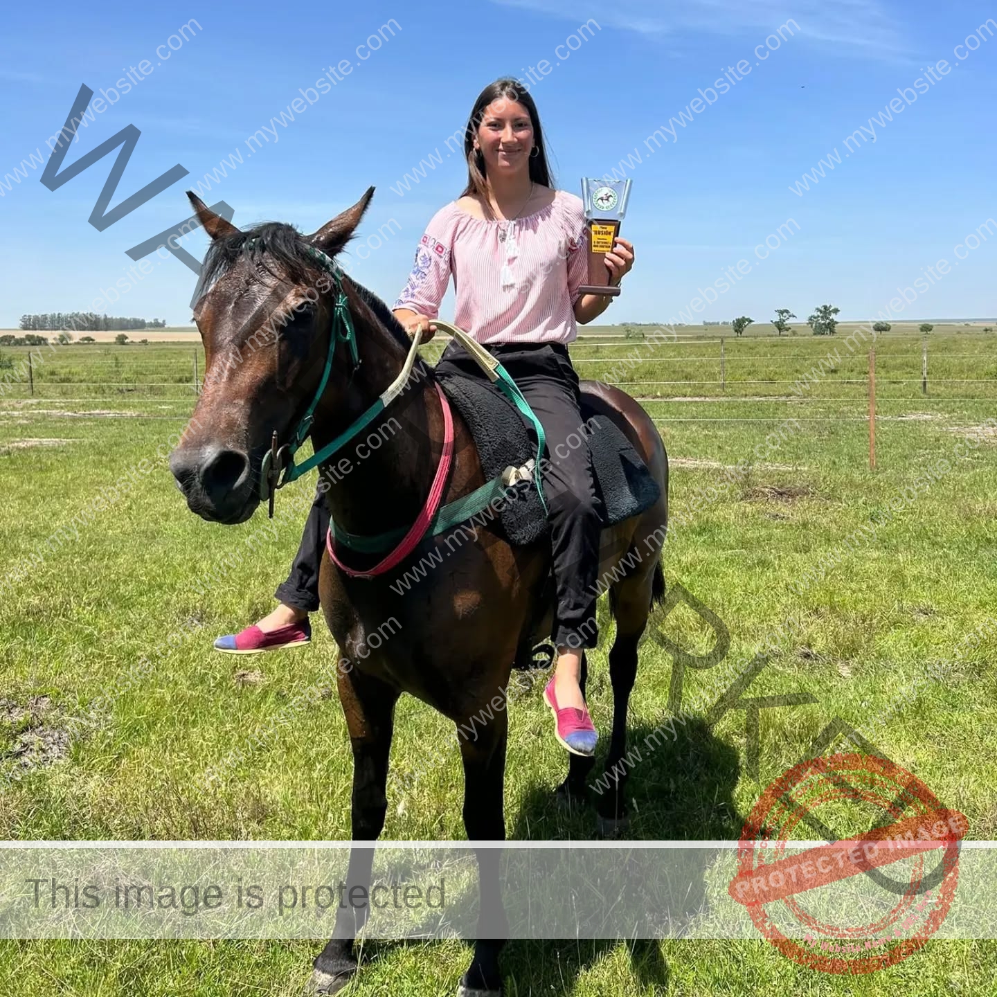 brandy-romero-uruguay-brandy_rom08-0033 Brandy Romero, a young woman, sits on a brown horse in grassy Uruguay, holding a trophy and smiling under the clear blue sky.