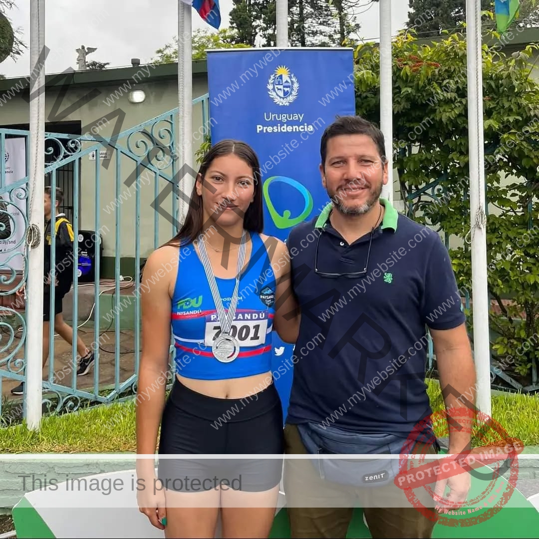 brandy-romero-uruguay-brandy_rom08-0046 Brandy Romero Track and field athlete Brandy Romero, wearing a medal, stands next to a smiling man before a Uruguay Presidencia banner outdoors.