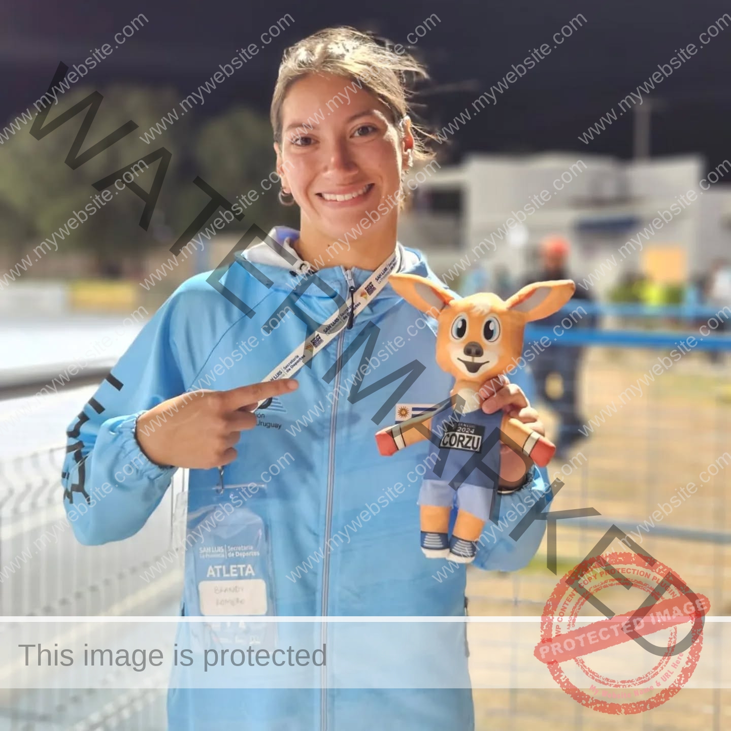 brandy-romero-uruguay-brandy_rom08-0065 Brandy Romero A smiling woman in a blue jacket holds a plush mascot and her ATLETA badge near a fence outdoors, track and field.