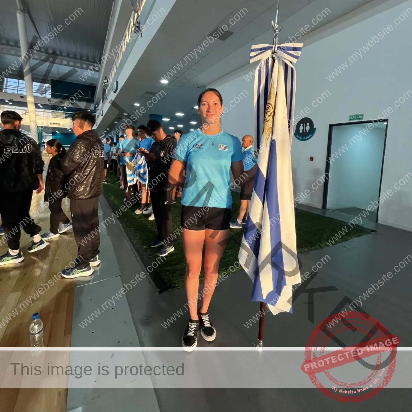 brandy-romero-uruguay-brandy_rom08-0072 Brandy Romero Brandy Romero, in a blue sports uniform, stands indoors holding a blue and white striped Uruguay flag; athletes line up behind.
