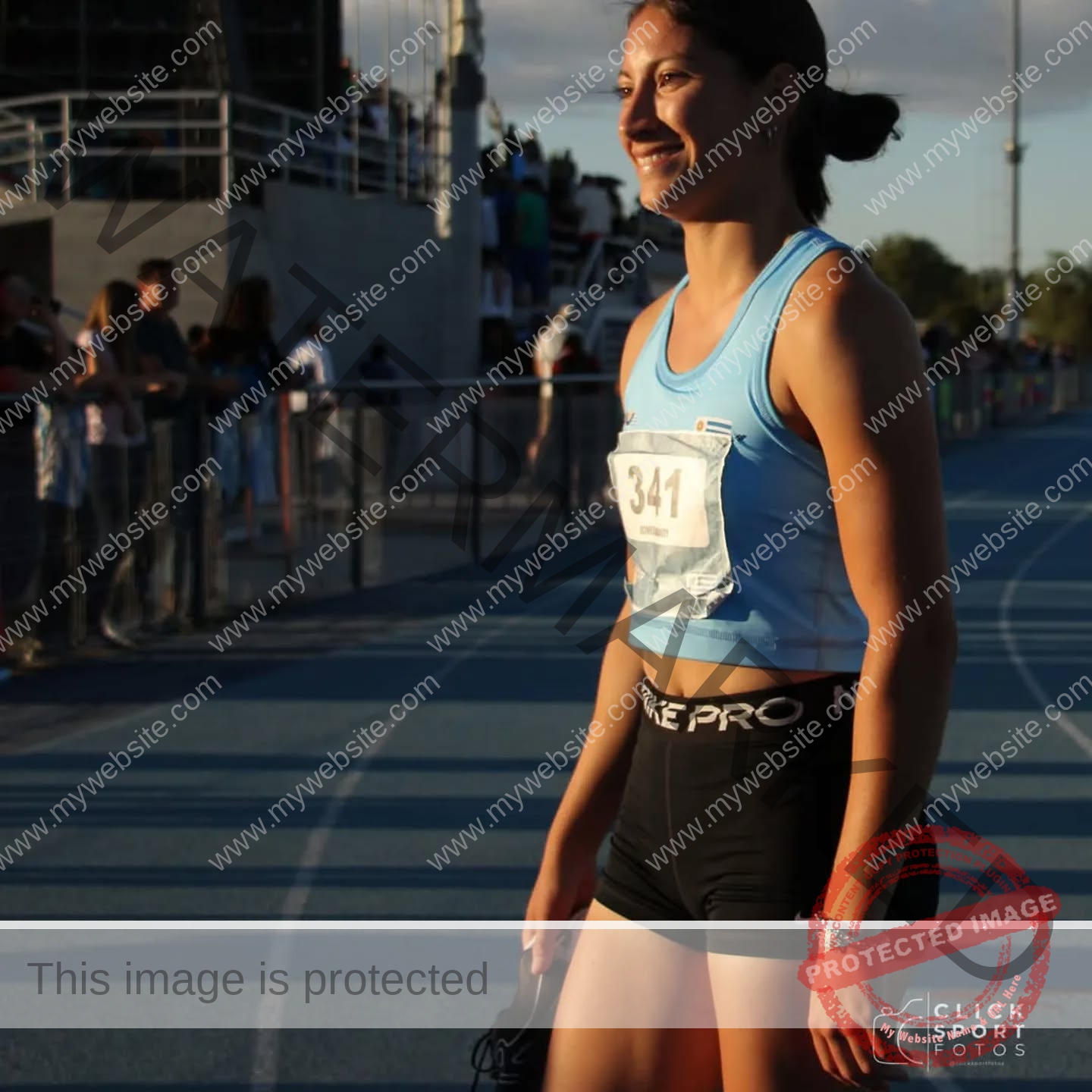 brandy-romero-uruguay-brandy_rom08-0075 Brandy Romero, a female track and field athlete in a blue top, black shorts, bib 341, smiles on a blue Uruguay track holding shoes.