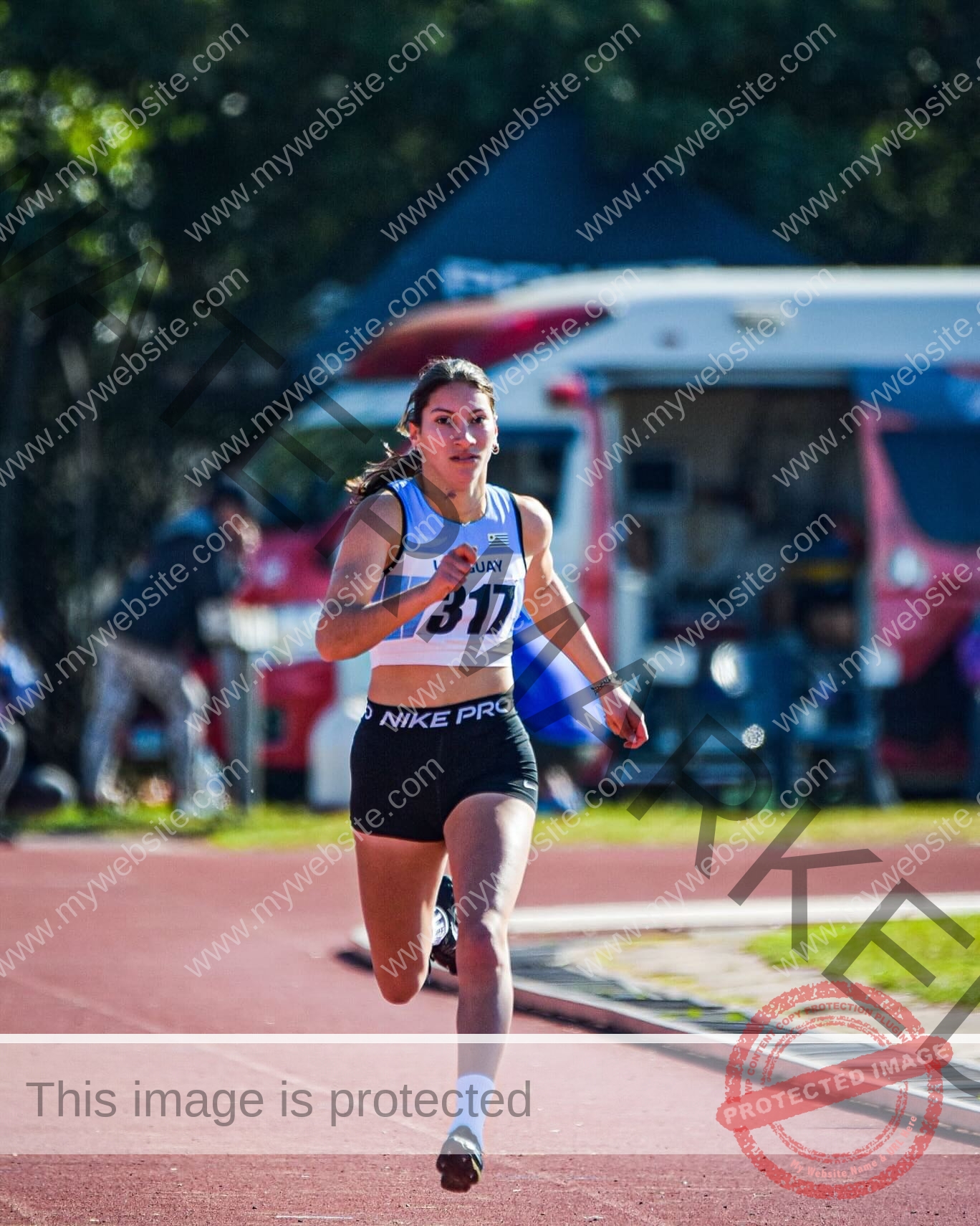 brandy-romero-uruguay-brandy_rom08-0088 Brandy Romero Brandy Romero, a female track and field athlete from Uruguay, wearing bib 317 and a blue-white top, sprints on an outdoor track with an ambulance and people blurred in the background.