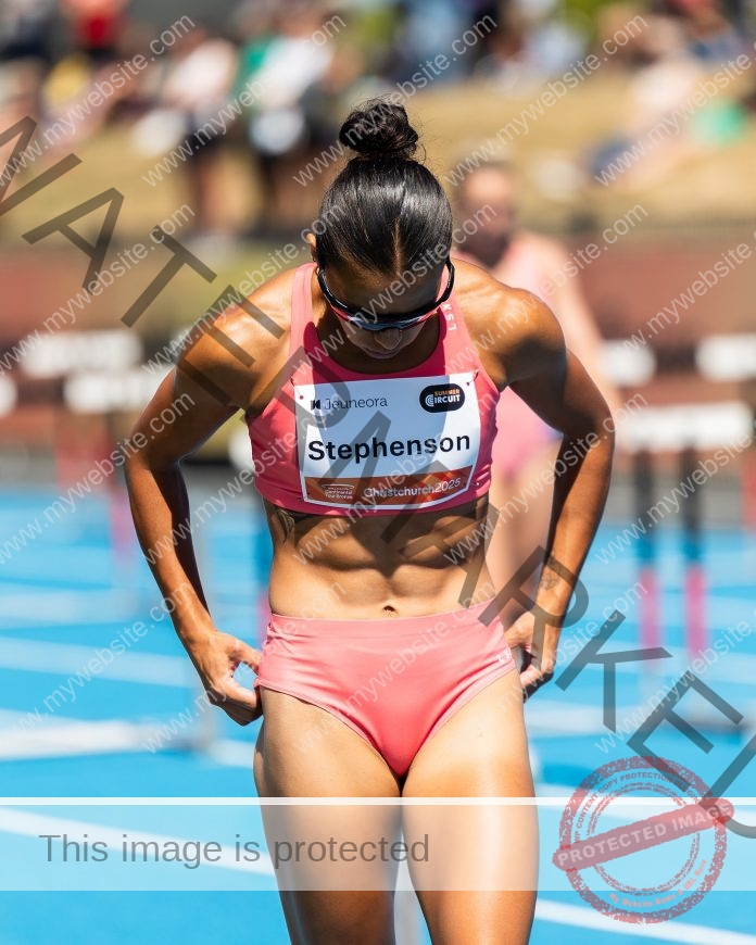 Briana Stephenson, a female track and field athlete in coral sportswear, stands on a blue track adjusting her shorts, with hurdles behind.