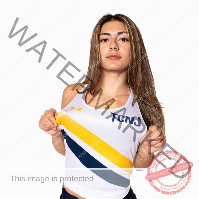 Cam Ruffini A young woman with long hair in a white TCNJ track tank top holds her shirt confidently, facing the camera on white.