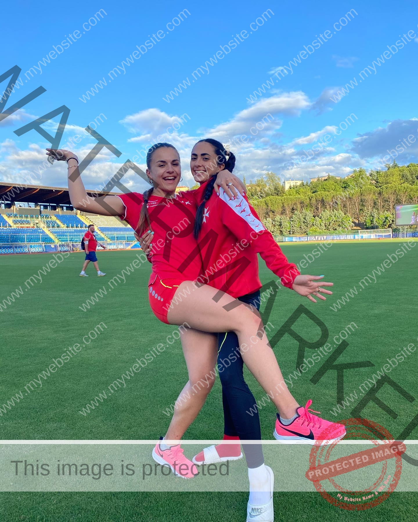 carla-scicluna-malta-carla.scicluna-01633 Carla Scicluna, track athlete from Malta: Two women in sporty red outfits smile and pose playfully on a grassy field with a stadium behind.