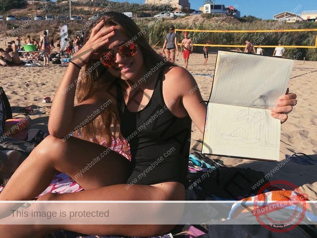 carla-scicluna-malta-carla.scicluna-01640 Carla Scicluna, track athlete from Malta, sits on a beach towel in sunglasses, showing a sketchbook with a figure drawing.