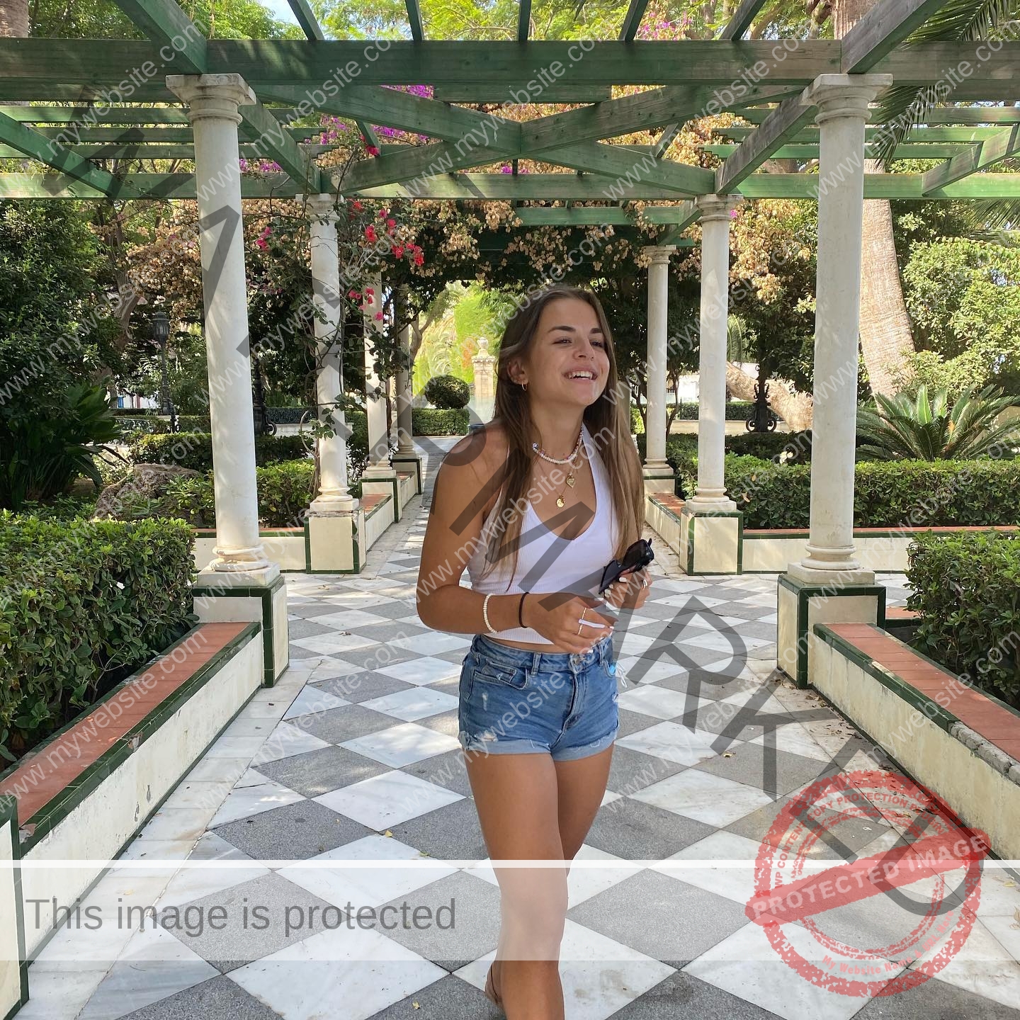 carla-scicluna-malta-carla.scicluna-01660 Carla Scicluna, track athlete from Malta, in a white tank top and shorts smiles under a pergola on a sunny checkered walkway.