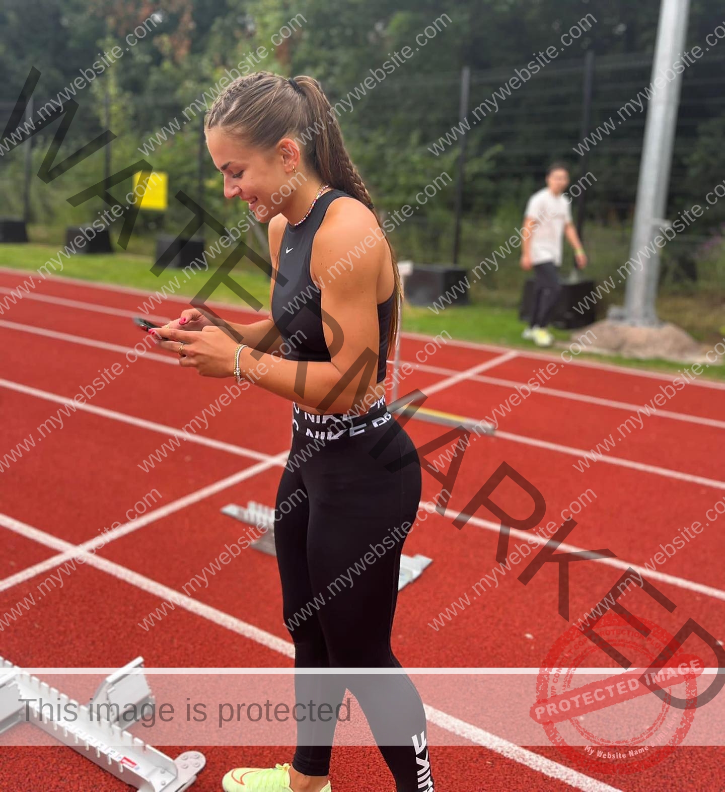 carla-scicluna-malta-carla.scicluna-01702 Carla Scicluna, track athlete from Malta, smiles at her phone on a red track with starting blocks, greenery, and another person nearby.