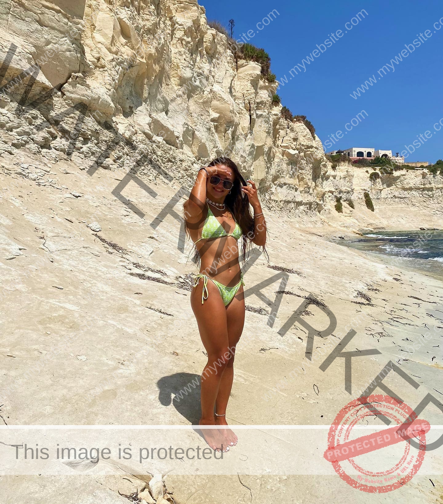 carla-scicluna-malta-carla.scicluna-01703 Carla Scicluna, track athlete from Malta, in a green bikini adjusts her sunglasses barefoot on a sandy beach near white cliffs.