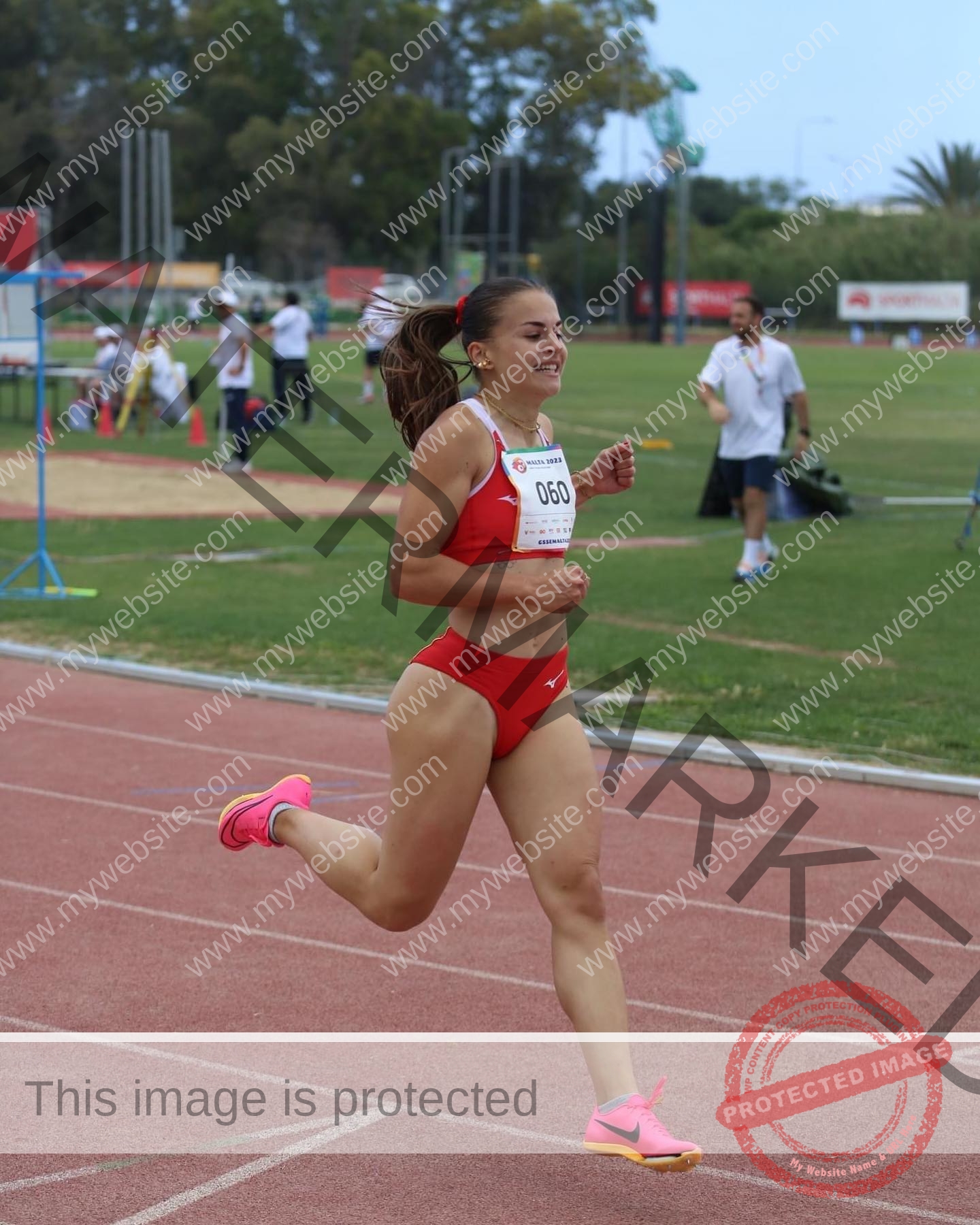 carla-scicluna-malta-carla.scicluna-01740 Carla Scicluna, track athlete from Malta, in a red sports outfit and pink shoes runs on a track with others and officials behind.