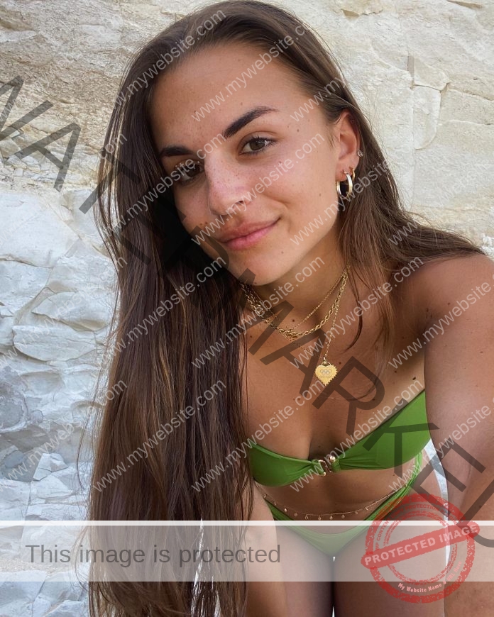 Carla Scicluna, track athlete from Malta, with long brown hair in a green bikini and gold jewelry poses before light rocks, smiling.