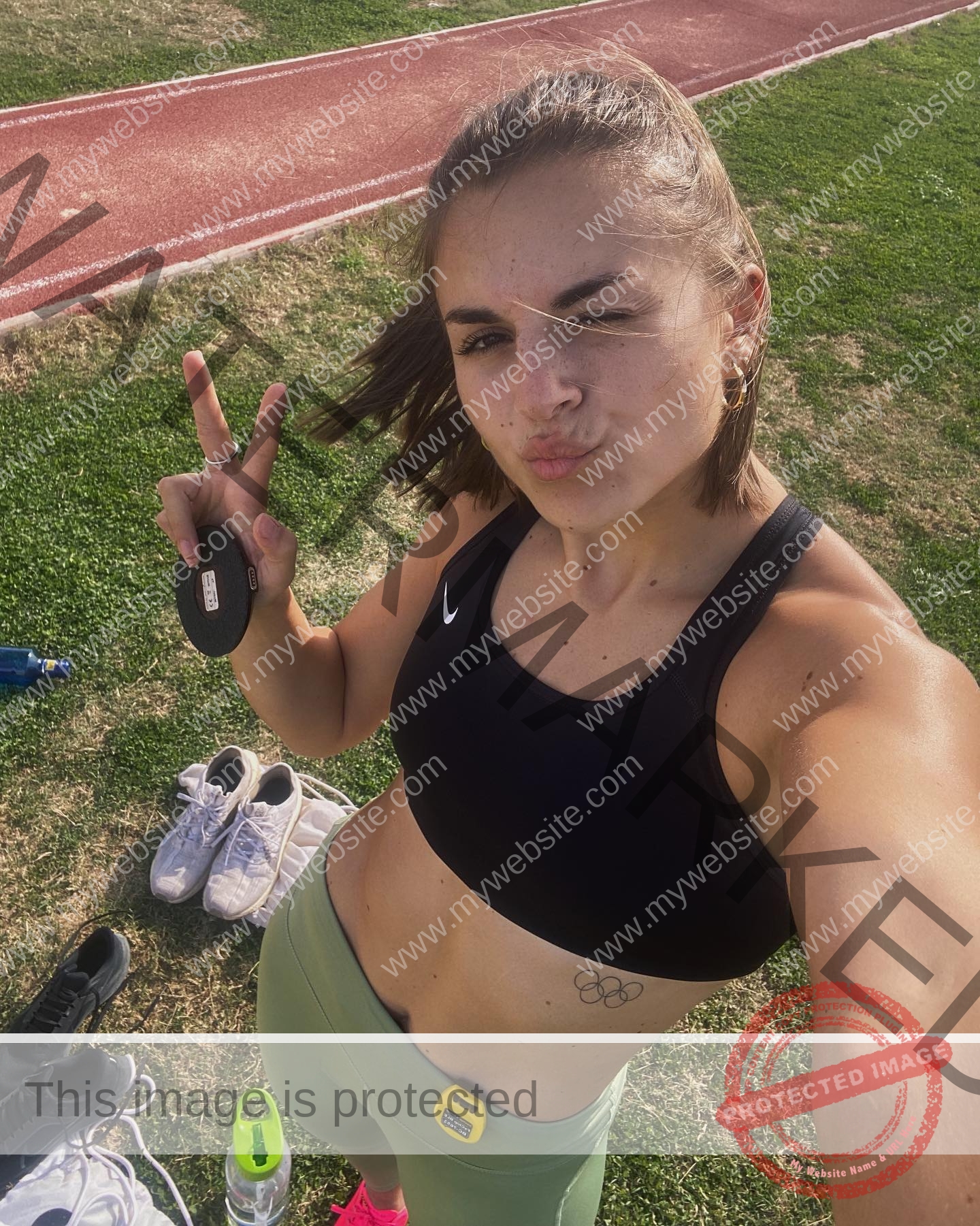 carla-scicluna-malta-carla.scicluna-01771 Carla Scicluna, track athlete from Malta, takes a selfie on a running track with a peace sign; workout gear sits nearby on the grass.
