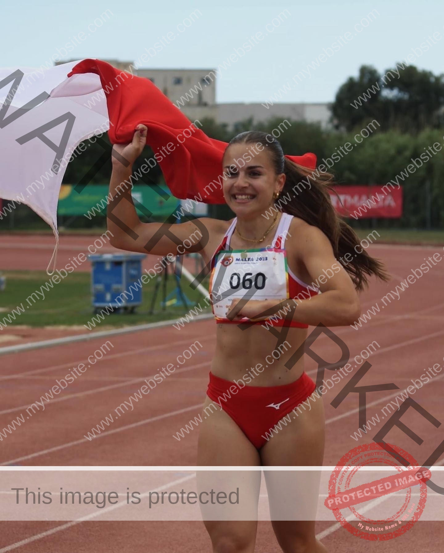 carla-scicluna-malta-carla.scicluna-01773 Carla Scicluna, track athlete from Malta, wears red sports gear and bib 060, running with a red and white flag raised in celebration.