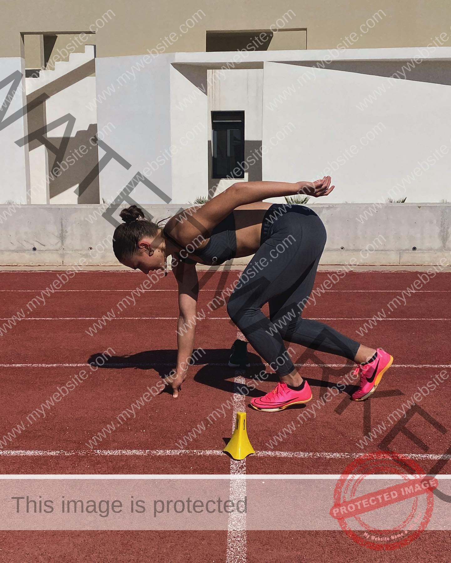 carla-scicluna-malta-carla.scicluna-01783 Carla Scicluna, track athlete from Malta, in athletic gear crouches at a starting line, poised to sprint with a yellow cone ahead.