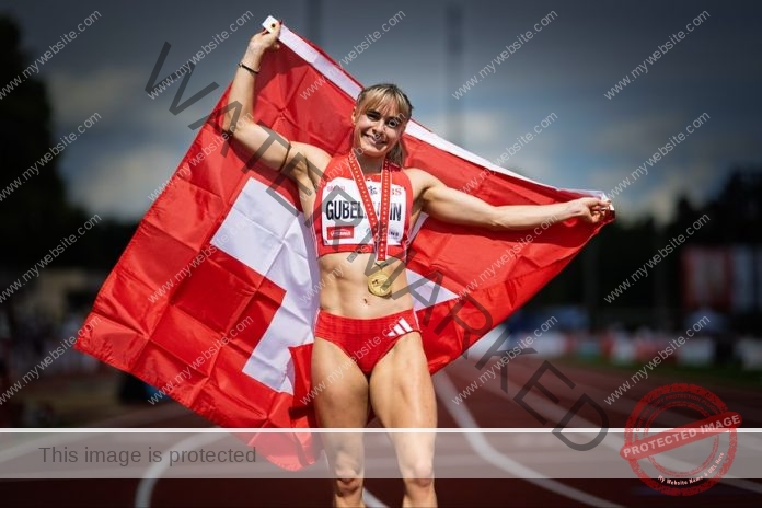 Catia Gubelmann, a female track and field athlete, stands on a Swiss track, smiling with a medal and flag, celebrating victory.