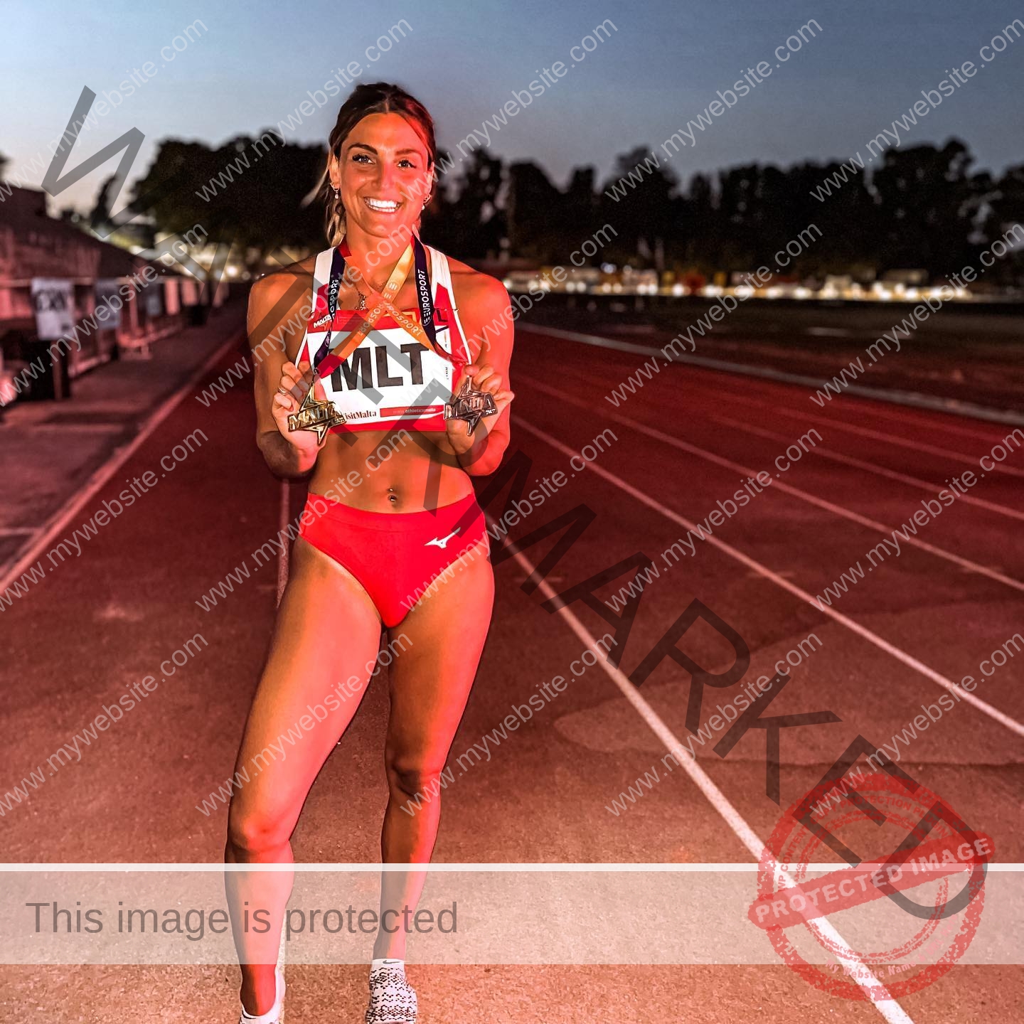 charlotte-wingfield-malta-charlottewingfield.oly-03737 Charlotte Wingfield, track athlete from Malta, in red uniform on a dimly lit track at dusk, smiling and holding her bib number.