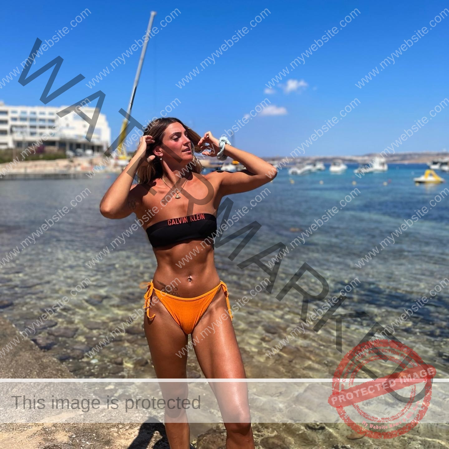 charlotte-wingfield-malta-charlottewingfield.oly-03745 Charlotte Wingfield, track athlete from Malta, stands on a rocky shore in a black and orange bikini by clear blue water, posing.