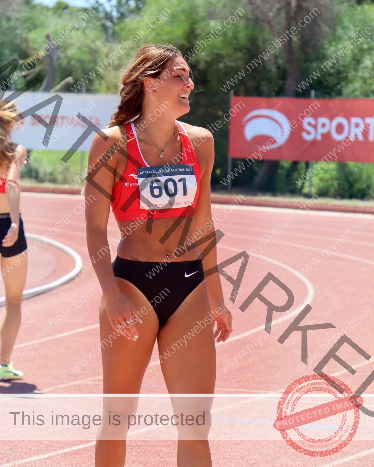 charlotte-wingfield-malta-charlottewingfield.oly-03769 Charlotte Wingfield, track athlete from Malta, smiles on a sunlit track wearing a red sports bra, black shorts, bib 601; trees behind.