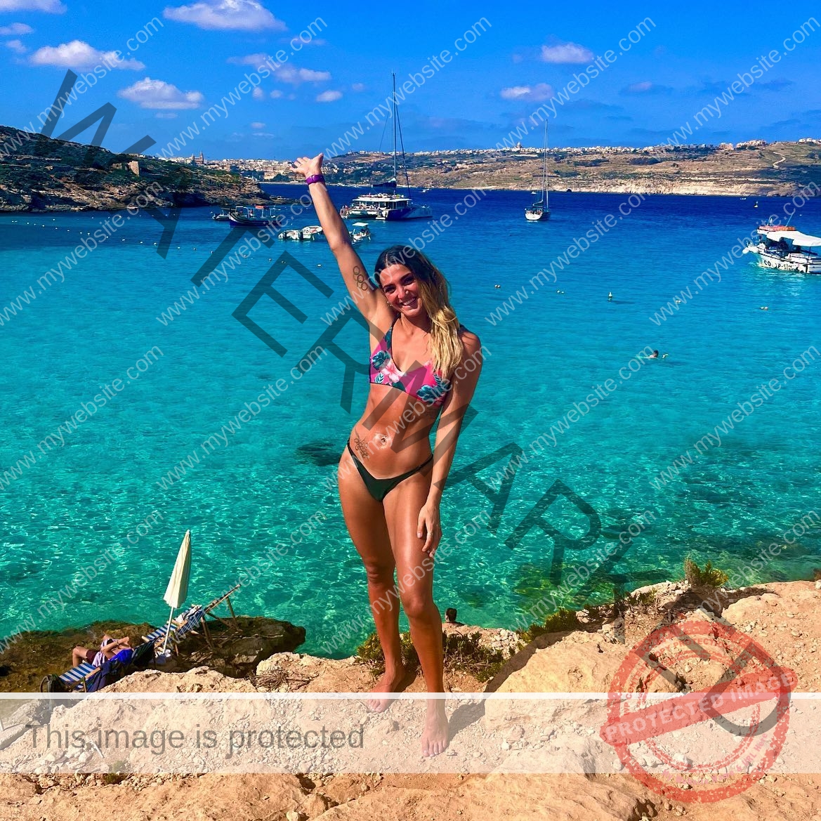 charlotte-wingfield-malta-charlottewingfield.oly-03790 Charlotte Wingfield, track athlete from Malta, stands smiling in a colorful bikini on rocks by turquoise water, arm raised, boats behind.