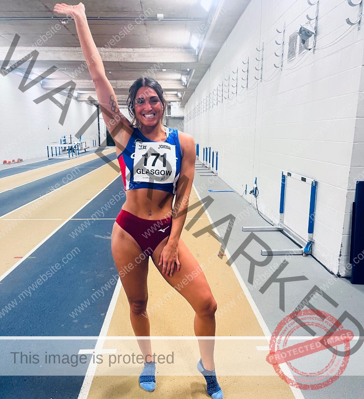 charlotte-wingfield-malta-charlottewingfield.oly-03817 Charlotte Wingfield, track athlete from Malta, in sportswear on an indoor track, holding up one arm and smiling. Bib 171 marked Glasgow.