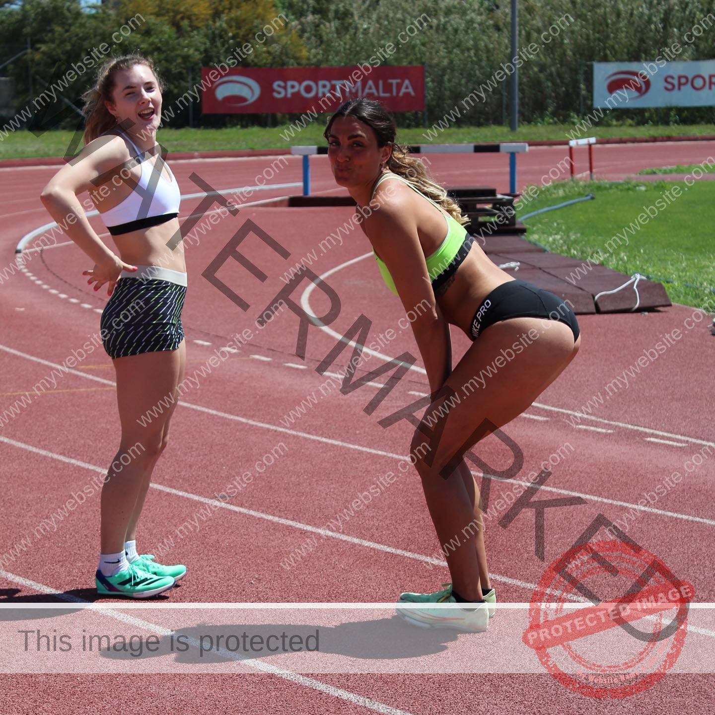 charlotte-wingfield-malta-charlottewingfield.oly-03832 Charlotte Wingfield, track athlete from Malta, poses playfully with another woman on a sunny red track, both smiling near SportMalta signs.
