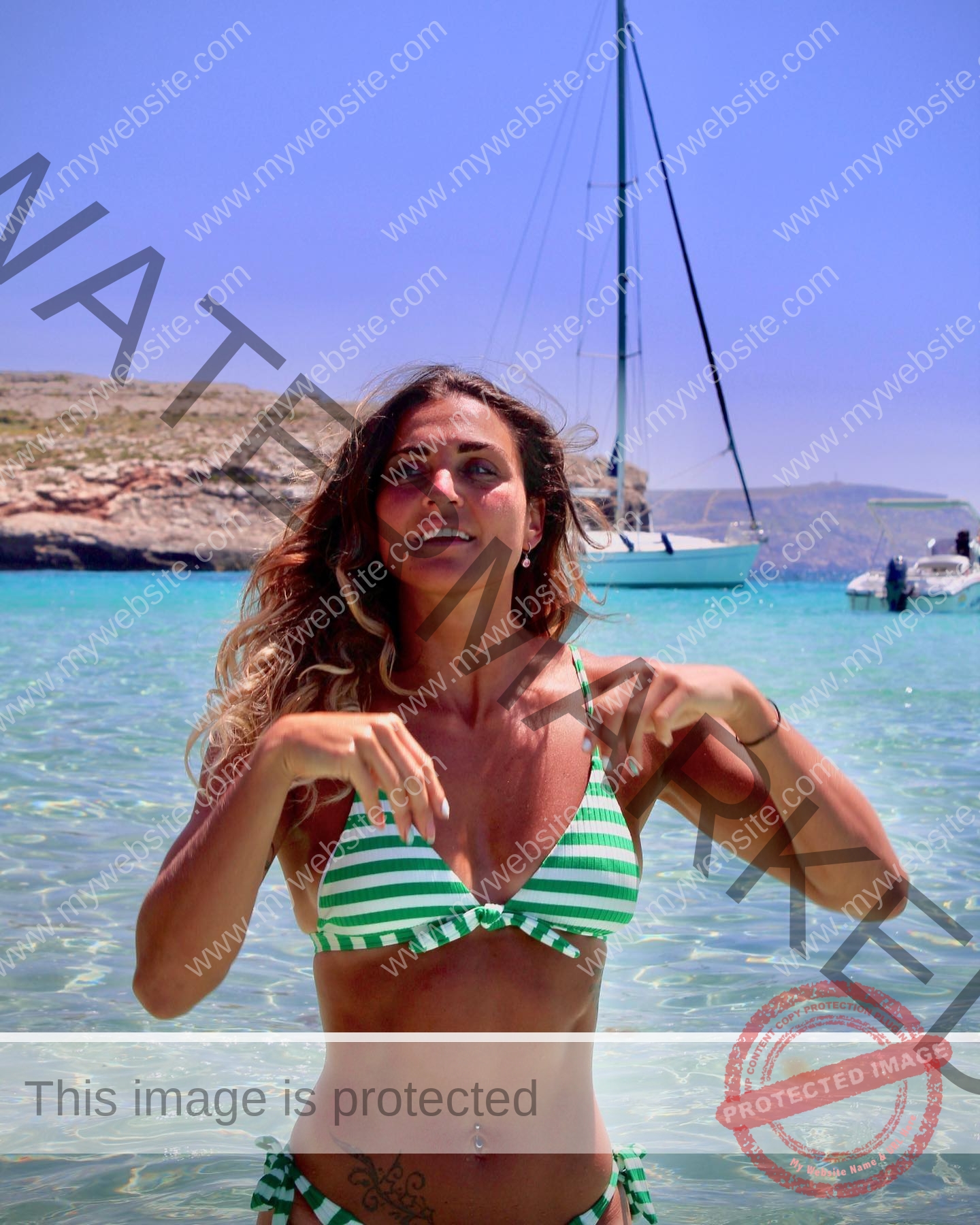 charlotte-wingfield-malta-charlottewingfield.oly-03841 Charlotte Wingfield, track athlete from Malta, in a green and white striped bikini stands in clear water with a sailboat and rocky shore.