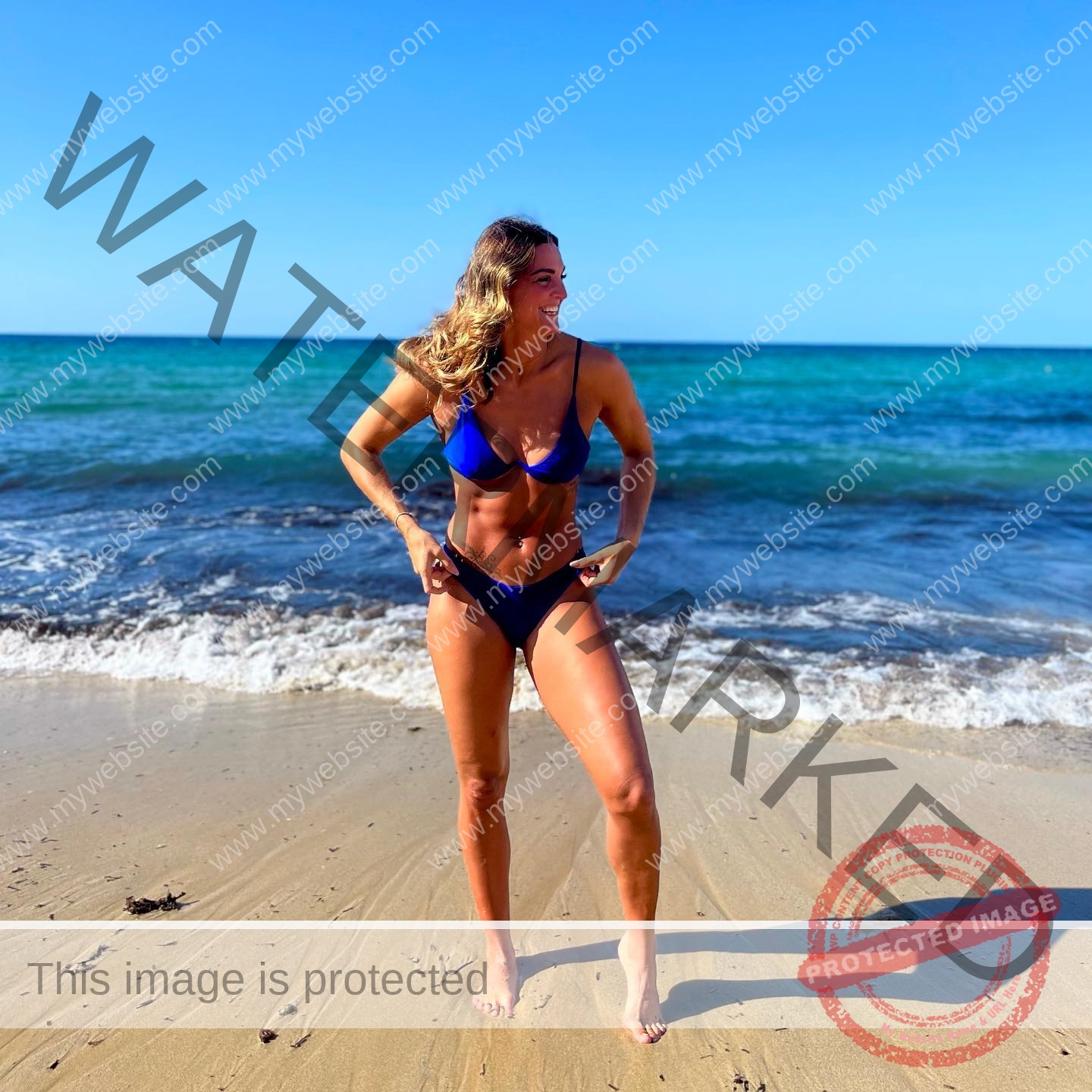 charlotte-wingfield-malta-charlottewingfield.oly-03919 Charlotte Wingfield, track athlete from Malta, in a blue bikini stands on a sunny sandy beach with waves behind her, smiling.