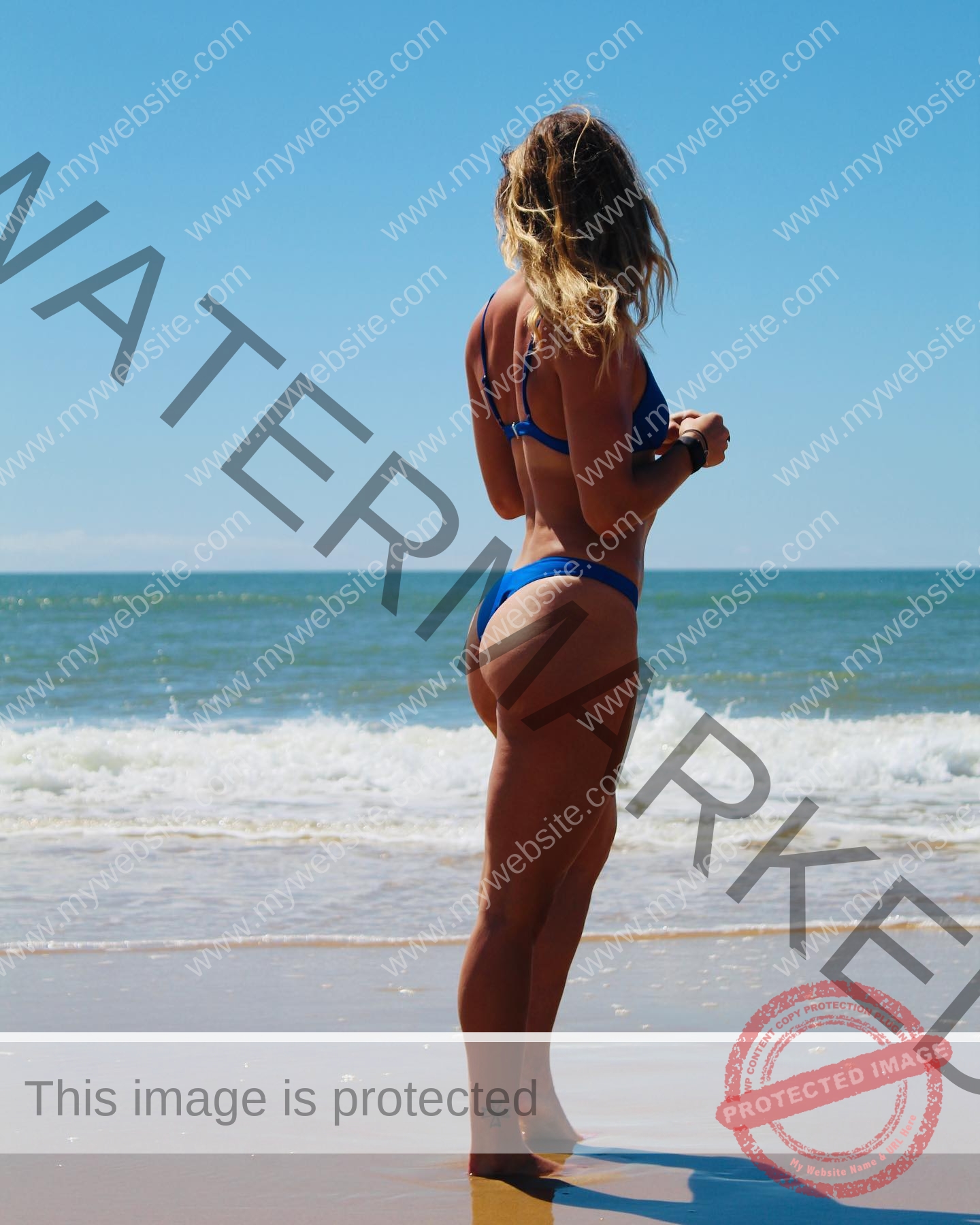 charlotte-wingfield-malta-charlottewingfield.oly-03931 Charlotte Wingfield, track athlete from Malta, in a blue bikini stands on a sandy beach, facing ocean waves under a clear blue sky.