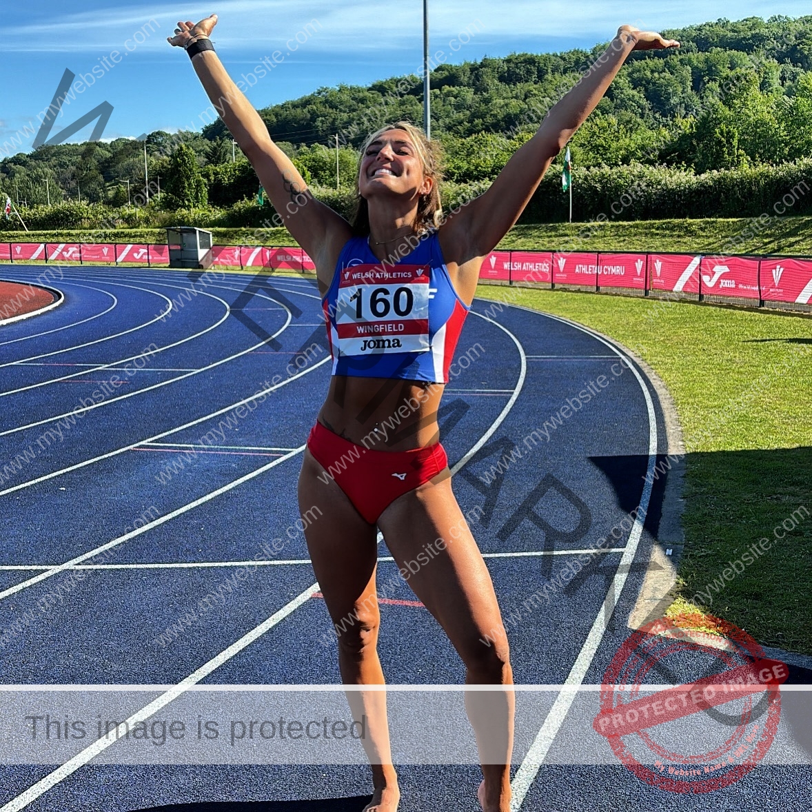 charlotte-wingfield-malta-charlottewingfield.oly-03941 Charlotte Wingfield, track athlete from Malta, in a red and blue uniform on a sunlit track, arms raised and smiling, bib 160 showing.