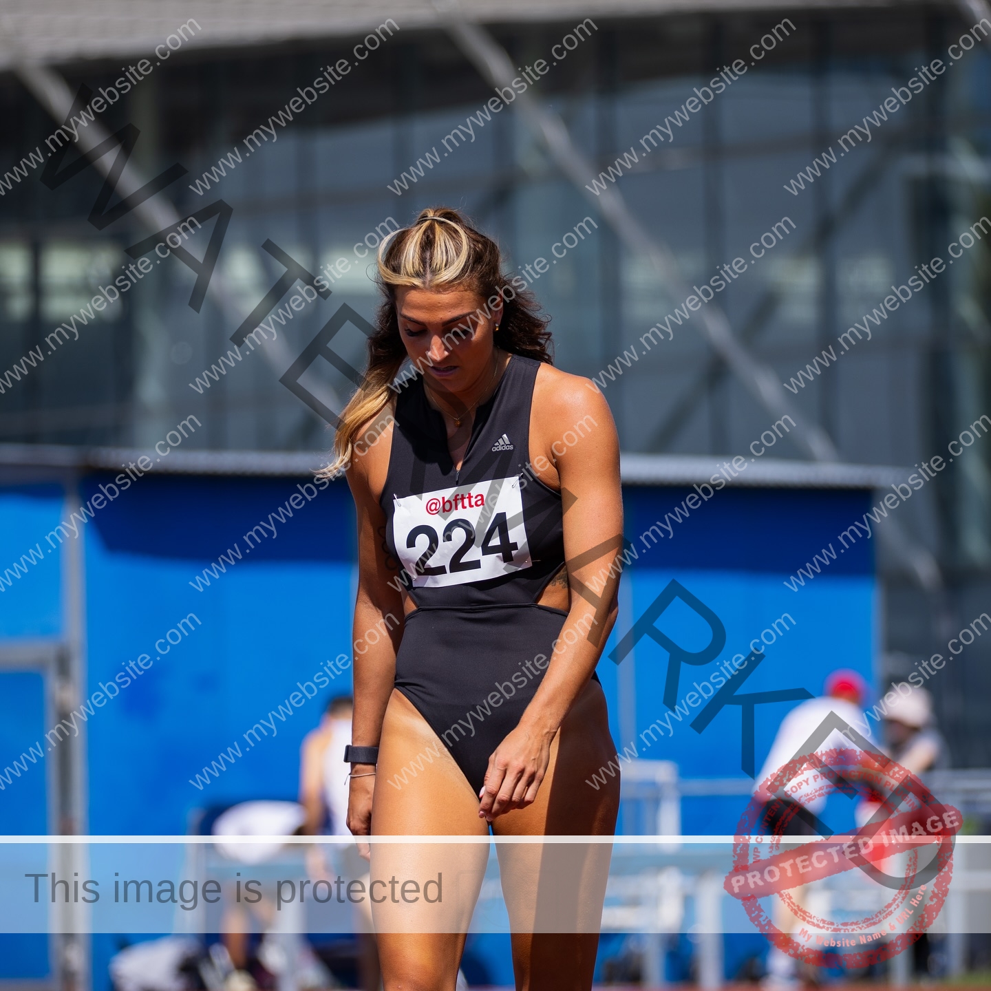 charlotte-wingfield-malta-charlottewingfield.oly-03947 Charlotte Wingfield, track athlete from Malta, in a black sports outfit and bib 224 stands on a track, looking down. Blue structure behind.