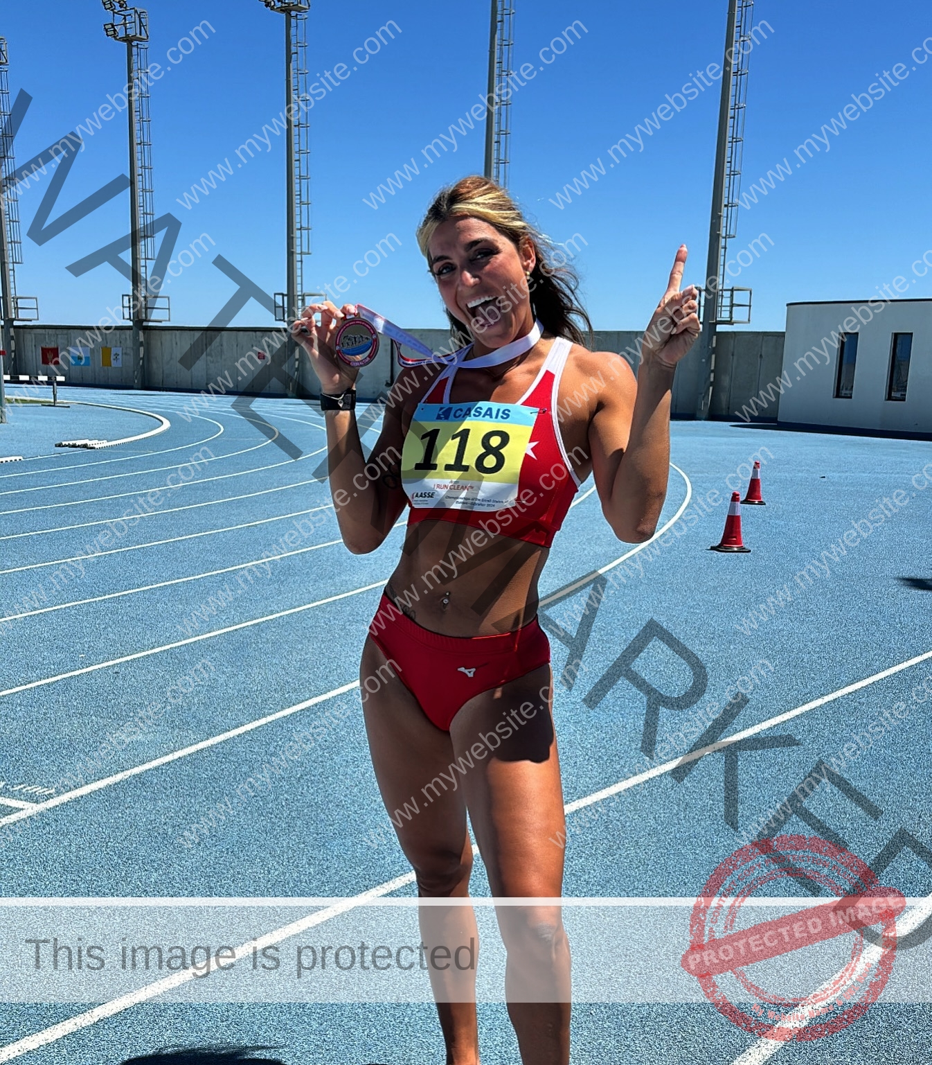 charlotte-wingfield-malta-charlottewingfield.oly-03976 Charlotte Wingfield, track athlete from Malta, in a red outfit on a blue track, holds a medal, smiling with finger raised—bib 118.