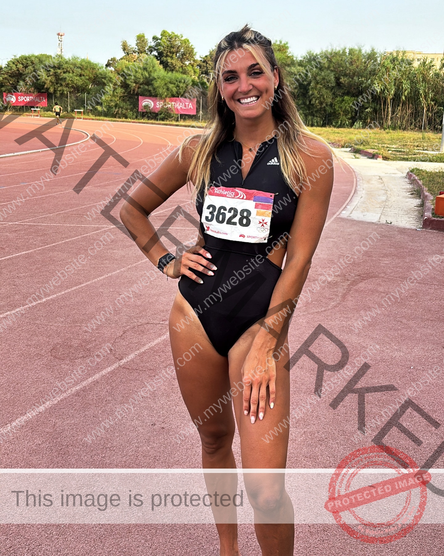 charlotte-wingfield-malta-charlottewingfield.oly-03977 Charlotte Wingfield, track athlete from Malta, smiling in a black Adidas outfit and bib 3628 on a track with SportMalta signs behind.