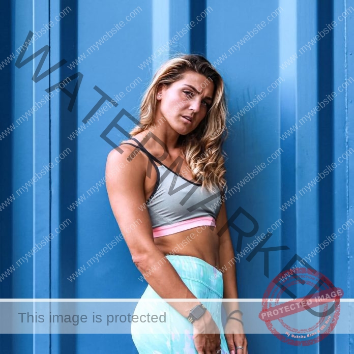 Charlotte Wingfield, track athlete from Malta, with wavy blonde hair in a grey sports bra and blue leggings stands by a blue metal wall.