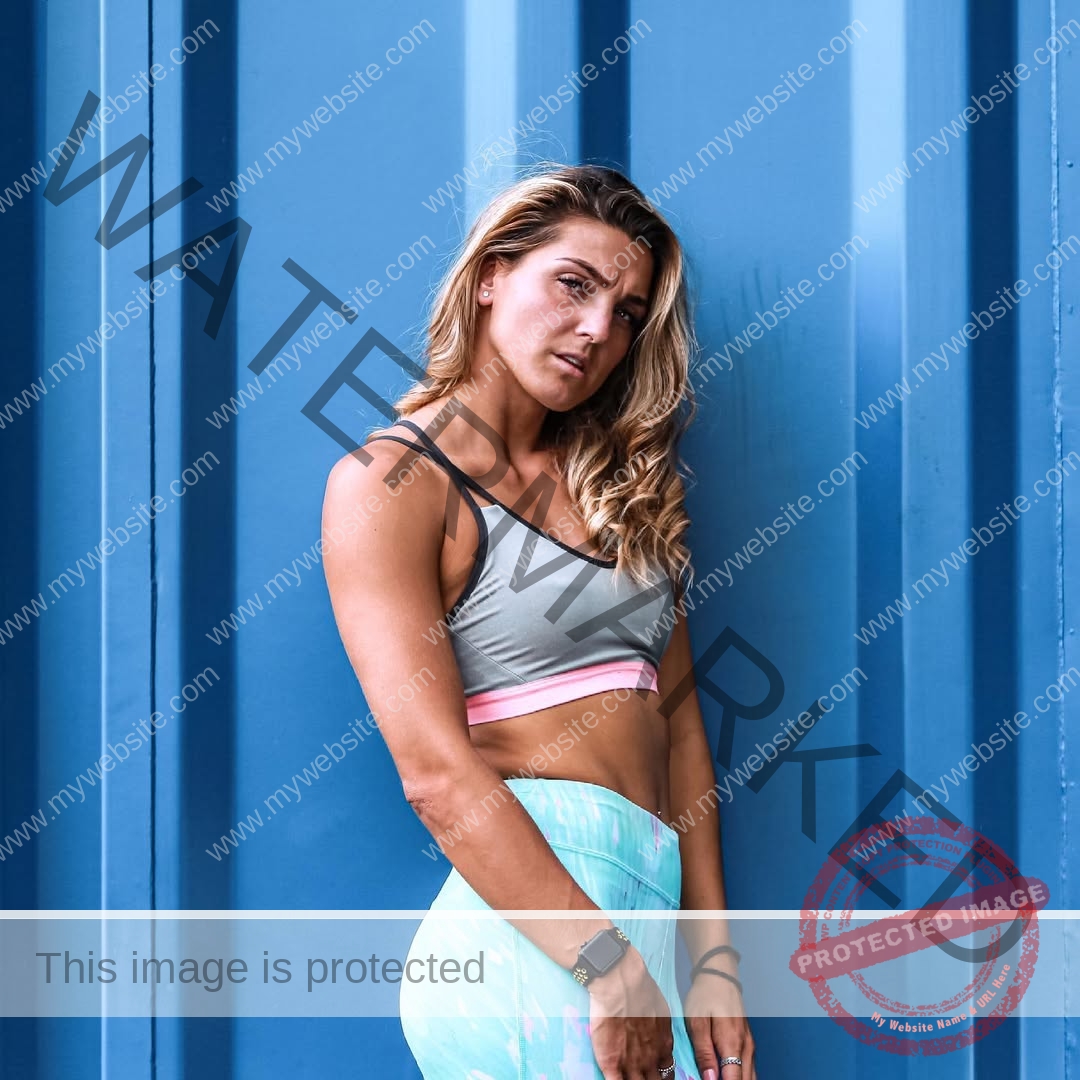 charlotte-wingfield-malta-charlottewingfield.oly-04261 Charlotte Wingfield, track athlete from Malta, with wavy blonde hair in a grey sports bra and blue leggings stands by a blue metal wall.