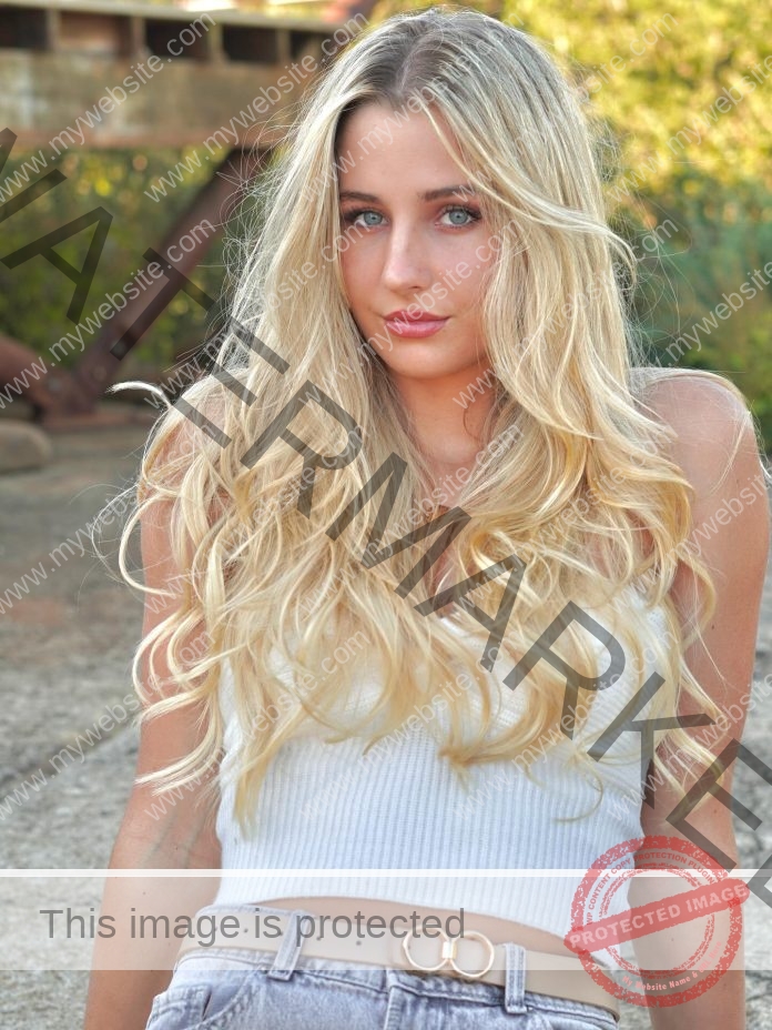 Charne Wansbury, track athlete from South Africa, with long blonde hair in a white tank and jeans, stands outdoors amid greenery.