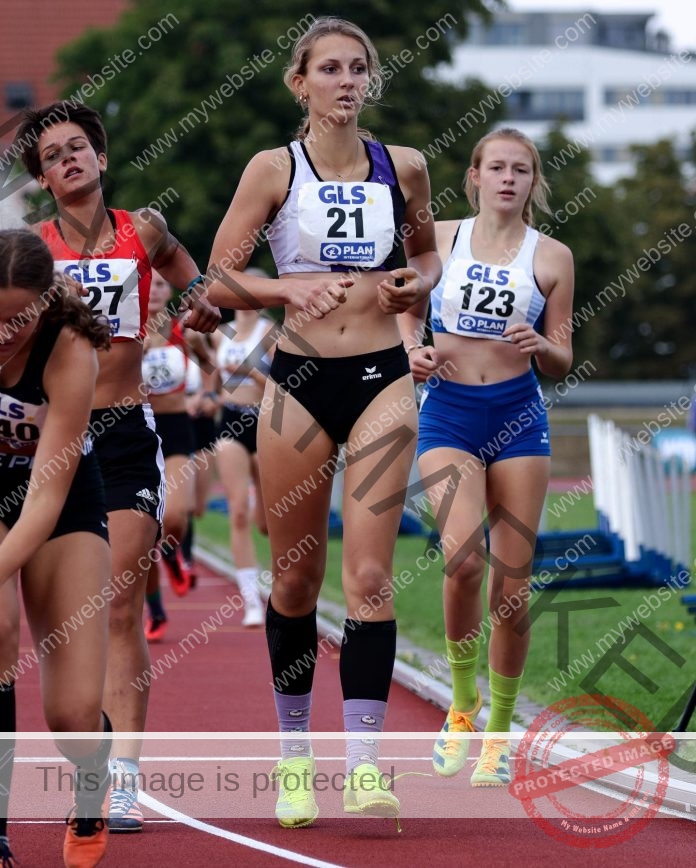 Chiara Wildner A group of young female track and field athletes, including Chiara Wildner in bib 21, race on a German track with trees and buildings behind.