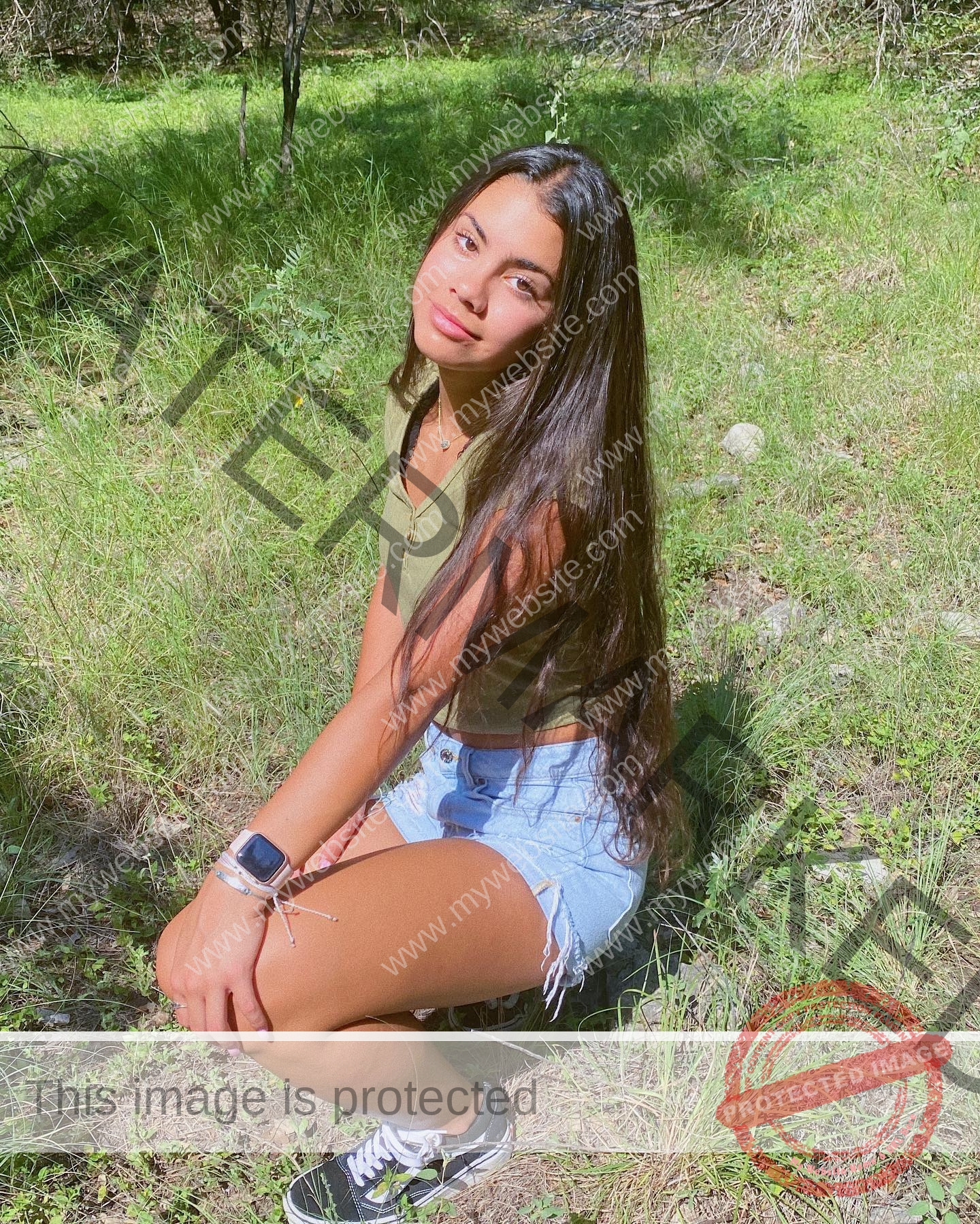 ciandra-carrico-trinity-ciandracarrico-3976 Ciandrad Carrico, track athlete from Trinity, with long dark hair, green top, denim shorts, kneels on grass looking up.