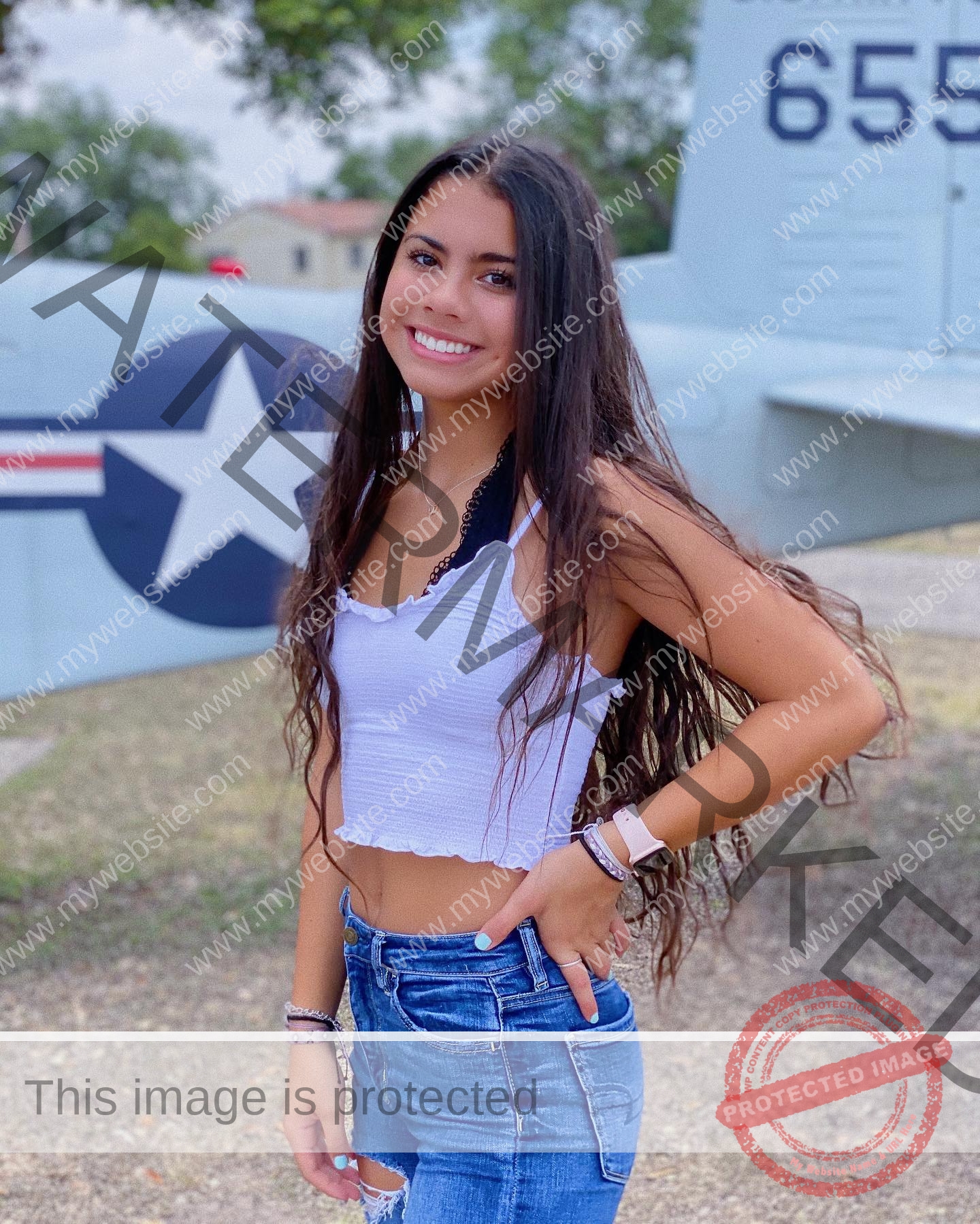ciandra-carrico-trinity-ciandracarrico-3983 Ciandra Carrico, track athlete from Trinity, with long brown hair, smiles in white crop top and jeans before a gray aircraft, star insignia.