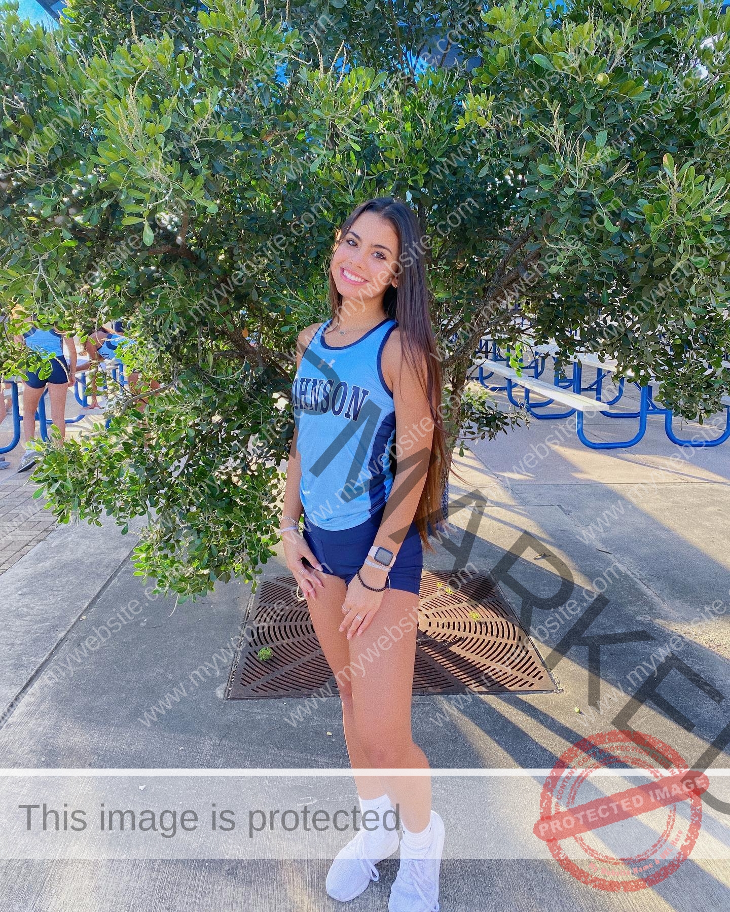 ciandra-carrico-trinity-ciandracarrico-4003 Ciandrad Carrico, track athlete from Trinity, in a blue Johnson uniform poses and smiles by a leafy tree on a sunny day.
