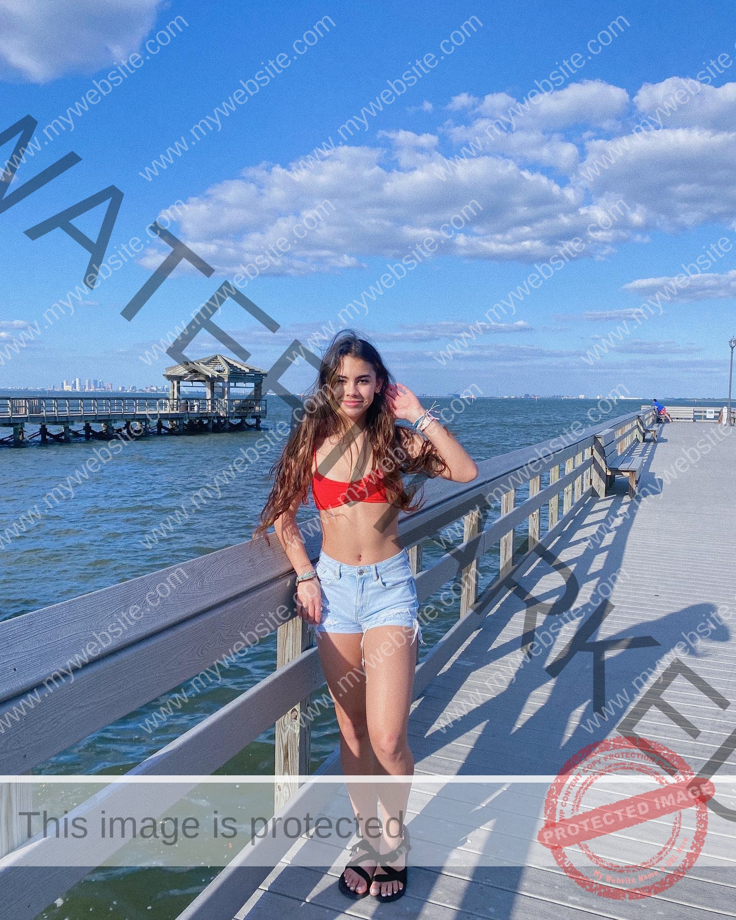 ciandra-carrico-trinity-ciandracarrico-4036 Ciandra Carrico, track athlete from Trinity, stands on a wooden pier in a red bikini top and denim shorts, smiling under a sunny sky.