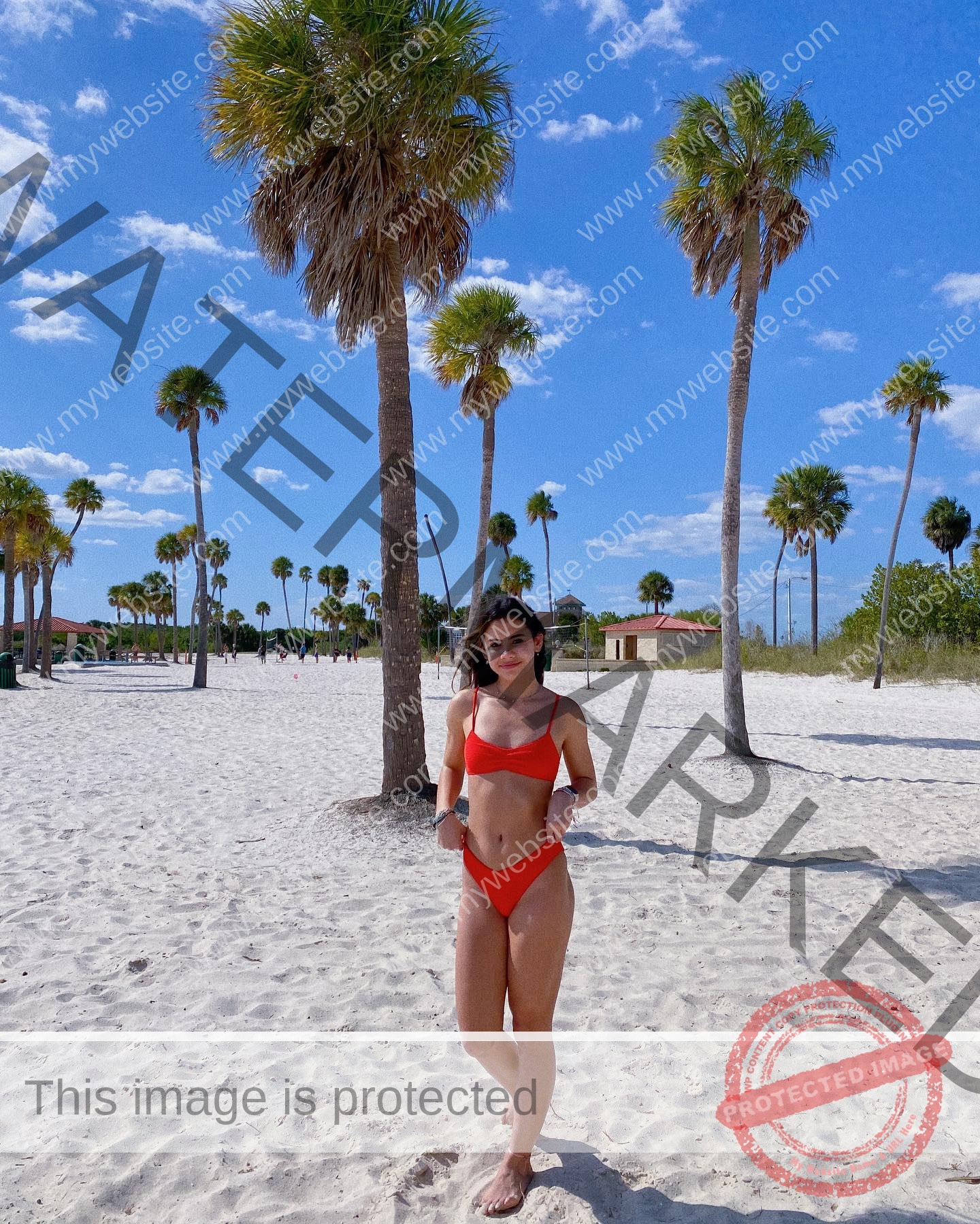 ciandra-carrico-trinity-ciandracarrico-4037 Ciandra Carrico, track athlete from Trinity, stands barefoot in a red bikini on a sandy, palm-lined beach under a blue sky.